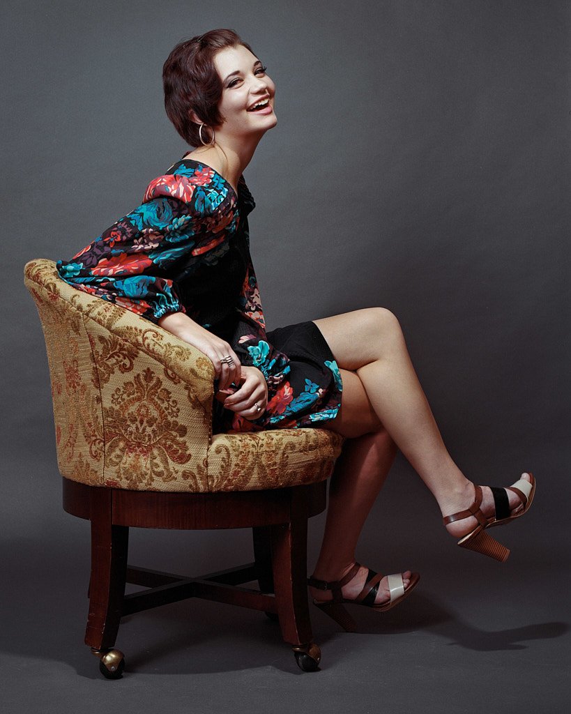 A smiling woman sitting sideways on a vintage patterned armchair against a dark gray background, wearing a colorful floral jacket, black shorts, and high-heeled sandals.