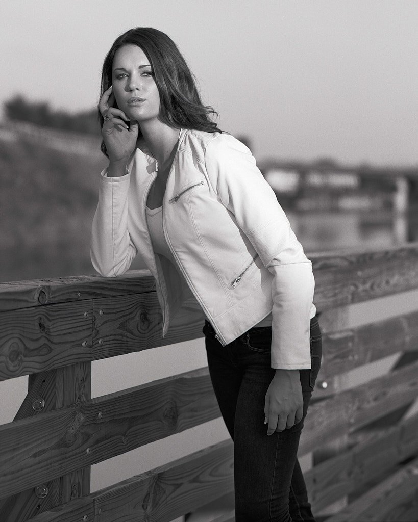 Black and white photo of a woman with wavy hair leaning on a wooden railing outdoors, wearing a white leather jacket and dark jeans. She has a thoughtful expression, with one hand resting on her face.