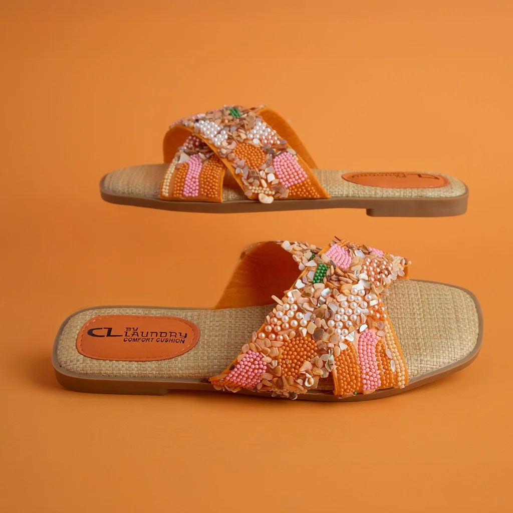 A pair of slide sandals with orange beaded and flower embellishments, labeled 'CLEAN BY LAUNDRY COMFORT CUSHION', against an orange background.