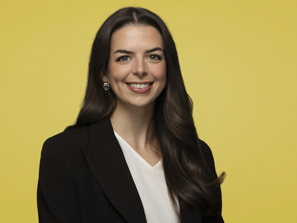 Professional headshot of a woman with long brown hair smiling, wearing a black blazer and a white top, against a yellow background.