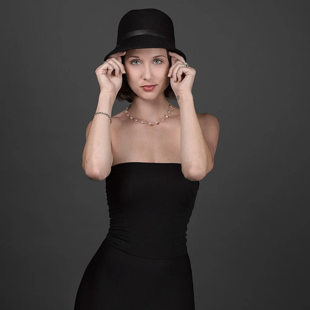 Woman wearing a black strapless dress and a black hat, holding the hat with both hands, against a gray background.