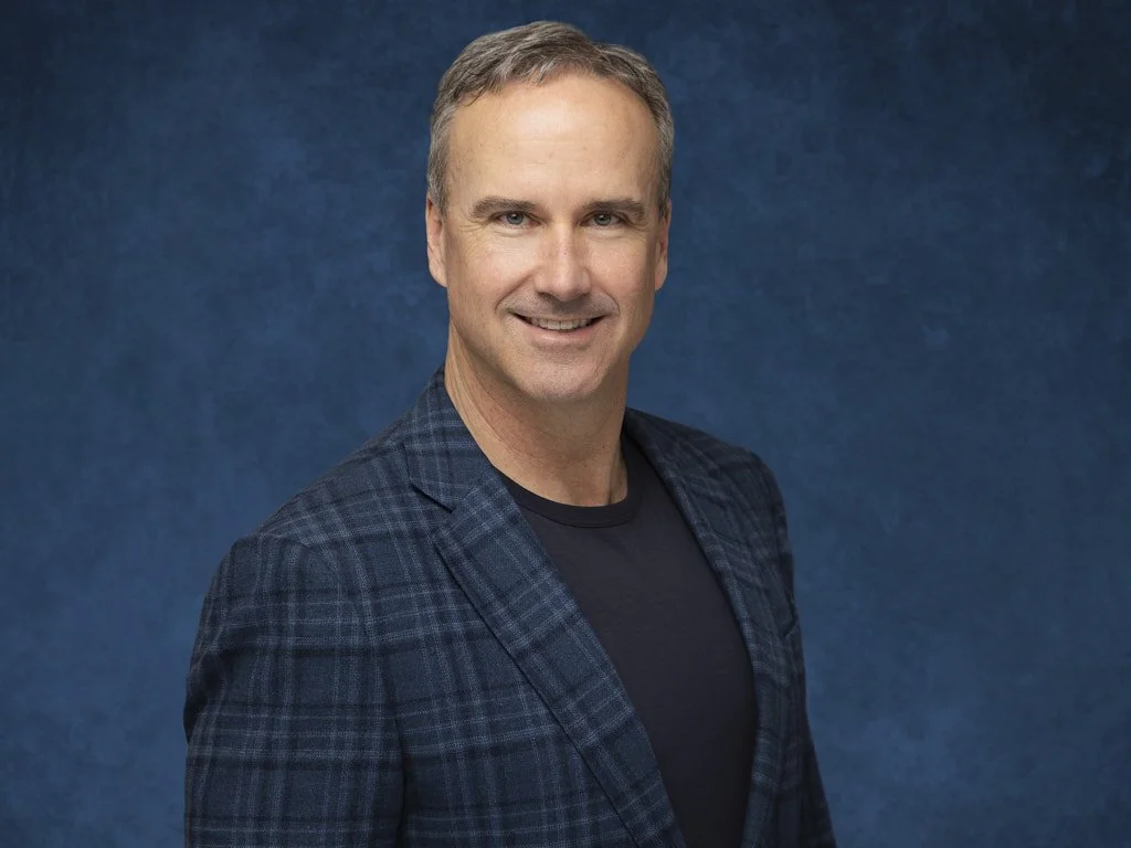 Professional headshot of a middle-aged man with short, graying hair smiling at the camera, wearing a dark checkered blazer over a black shirt, against a textured blue background.