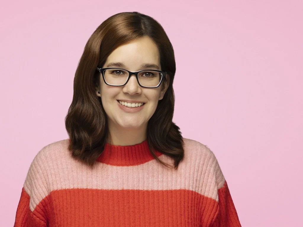 Headshot of a woman with brown hair wearing glasses and a pink and red sweater, smiling against a pink background.