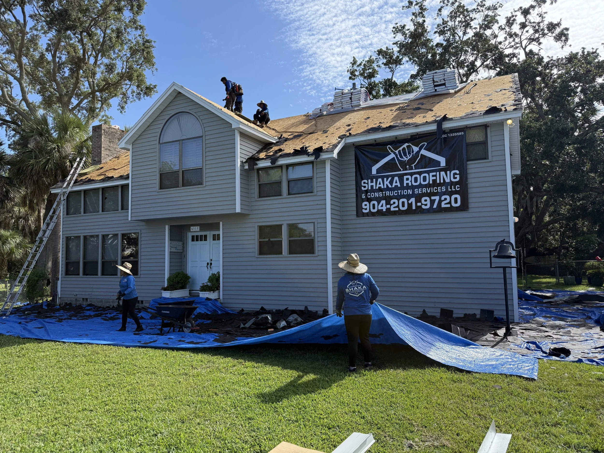 Workers repairing and replacing the roofing on a two-story house with gray siding, a front door, and multiple windows, with a large sign for Shaka Roofing & Construction Services LLC on the side of the house. The workers are on the roof and ground, w