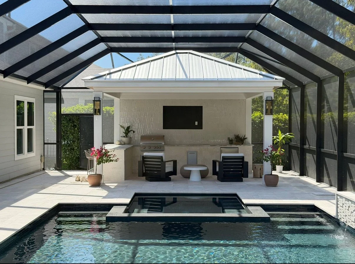 A screened-in outdoor patio with a pool in the foreground, seating area, outdoor kitchen with a grill, and wall-mounted TV, surrounded by potted plants and greenery.