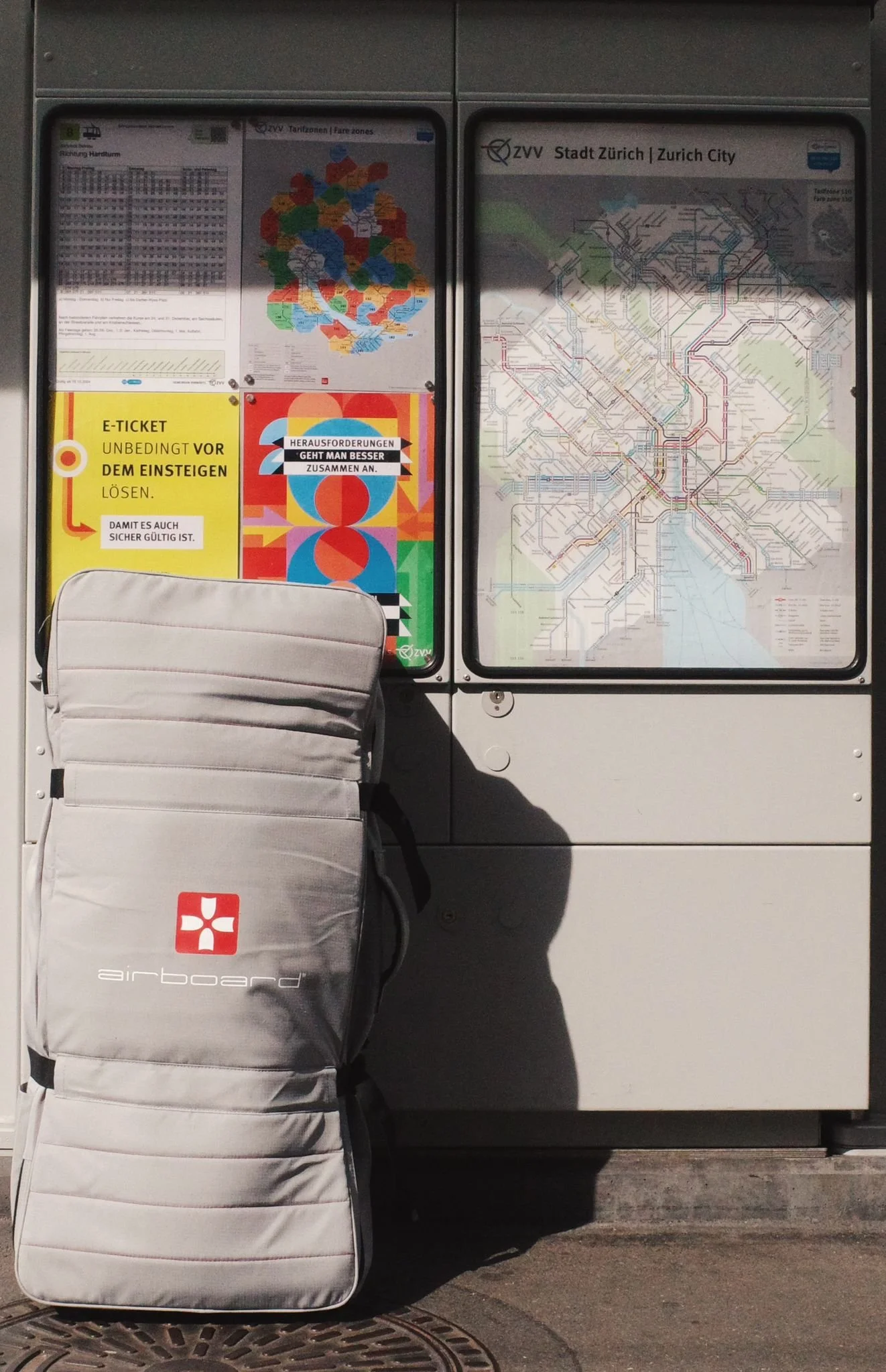 A travel backpack labeled 'airboard' leaning against a Zurich city transit information board with maps and schedules.