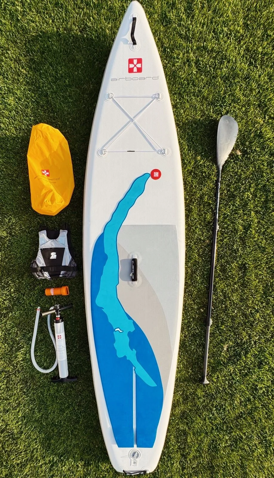 Inflatable stand-up paddleboard with a paddle, pump, small bag, and yellow dry bag on grass.