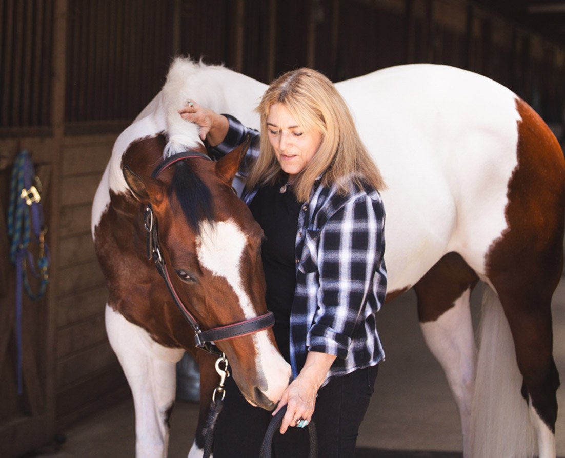 Cherie wears a black plaid shirt as she strokes the nose of a skewbald horse