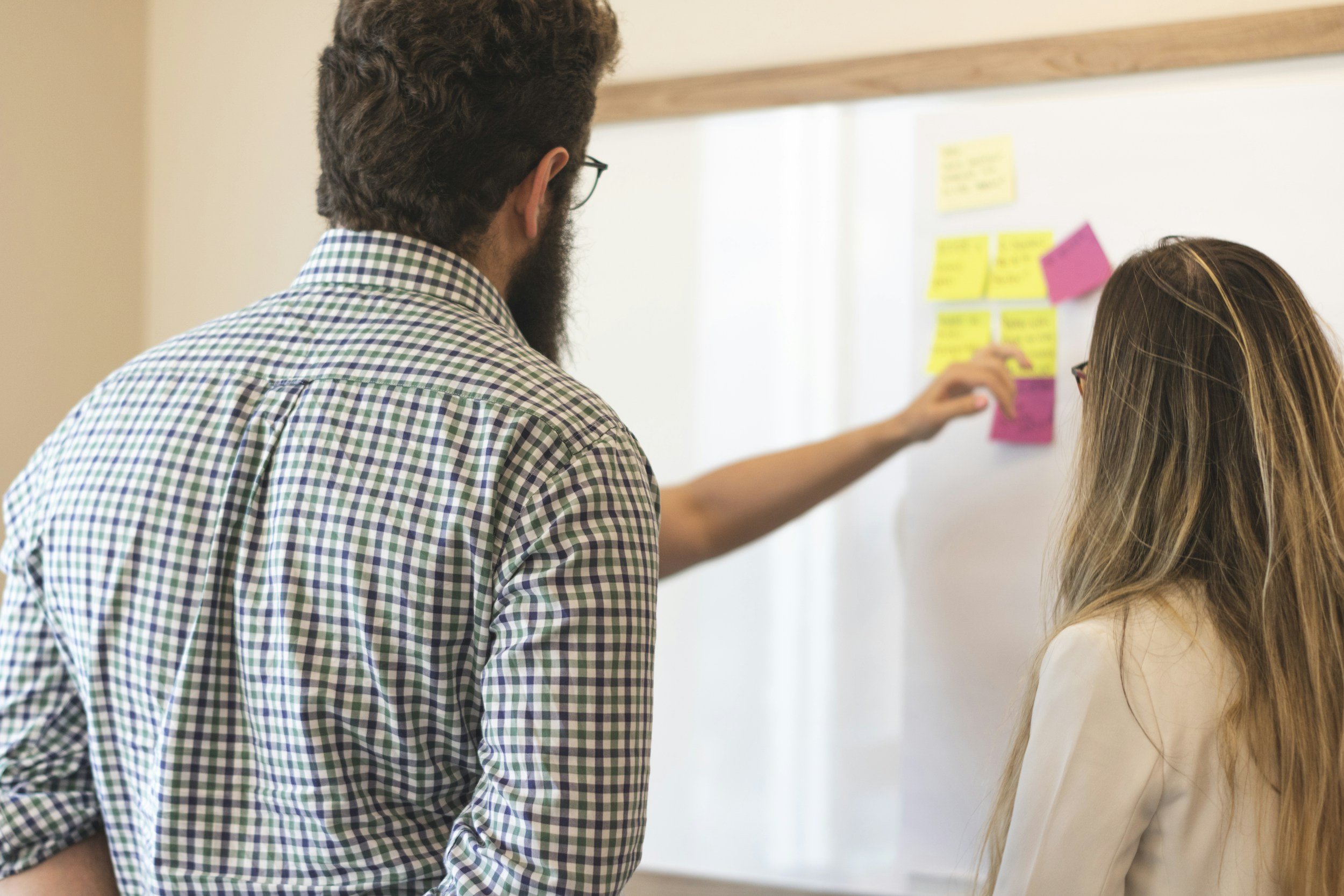 Two individuals, a man with glasses and a woman with long hair, are standing in front of a whiteboard with colorful sticky notes arranged on it. The man is pointing at the notes, and they seem to be discussing or collaborating on a project.