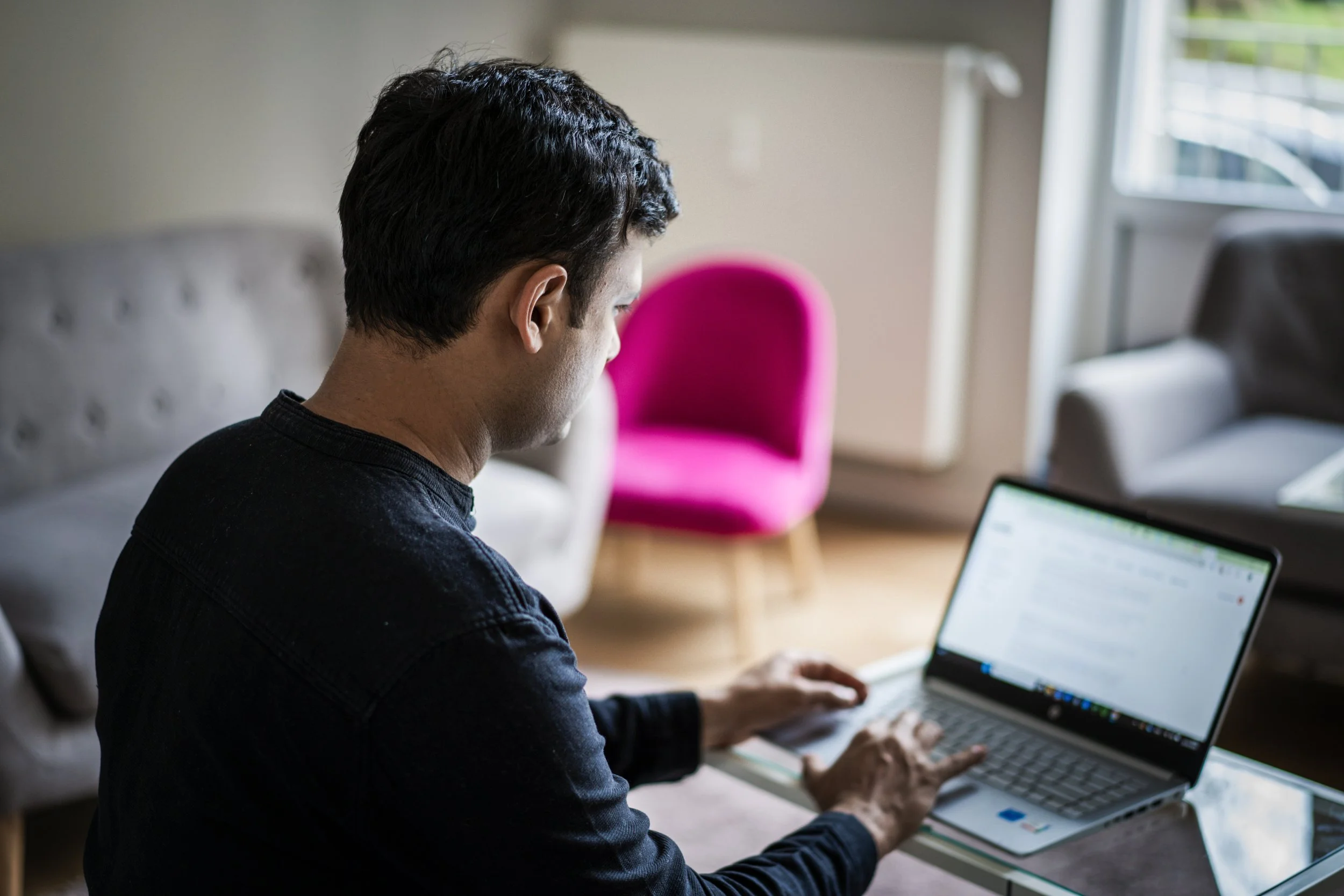 A man working on a laptop at a table in a living room with a pink chair and gray sofas in the background.