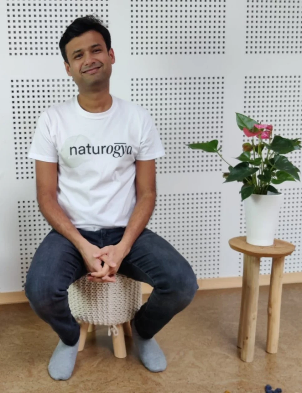 A man sitting on a small woven stool, wearing a white t-shirt with 'naturogya' printed on it, grey socks, and dark jeans, smiling at the camera. There is a potted plant with pink flowers on a wooden stand to his right, and a white perforated wall behind him.