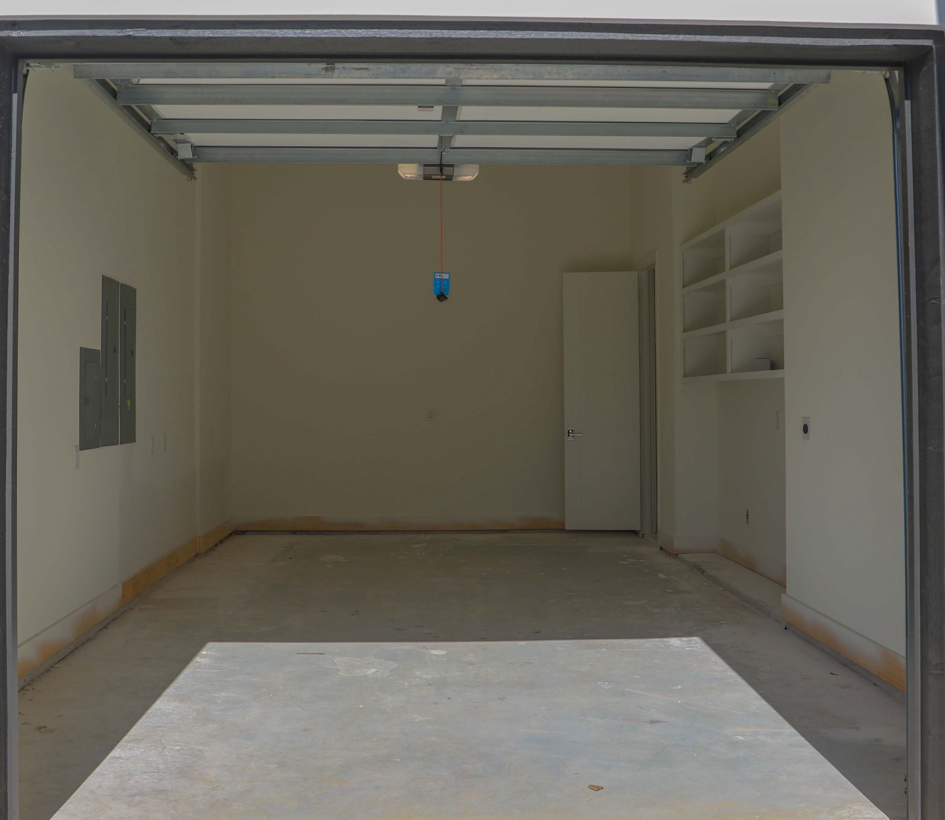 Empty garage with open door, exposed concrete floor, wall-mounted shelves, electrical panel, and overhead light fixture.