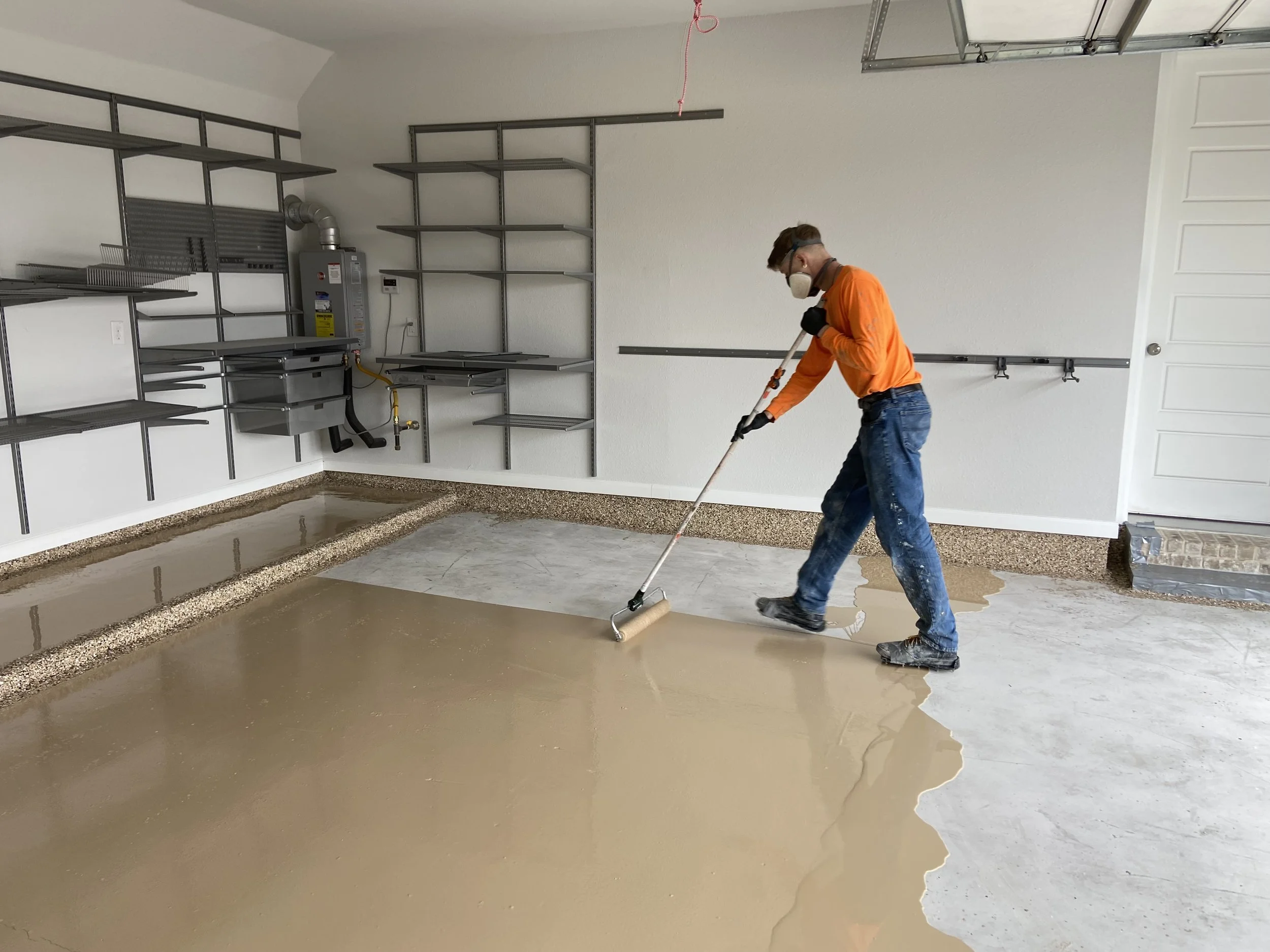 A person applying epoxy coating to a garage floor with a roller, wearing a mask and gloves. Shelving units are installed on the walls.