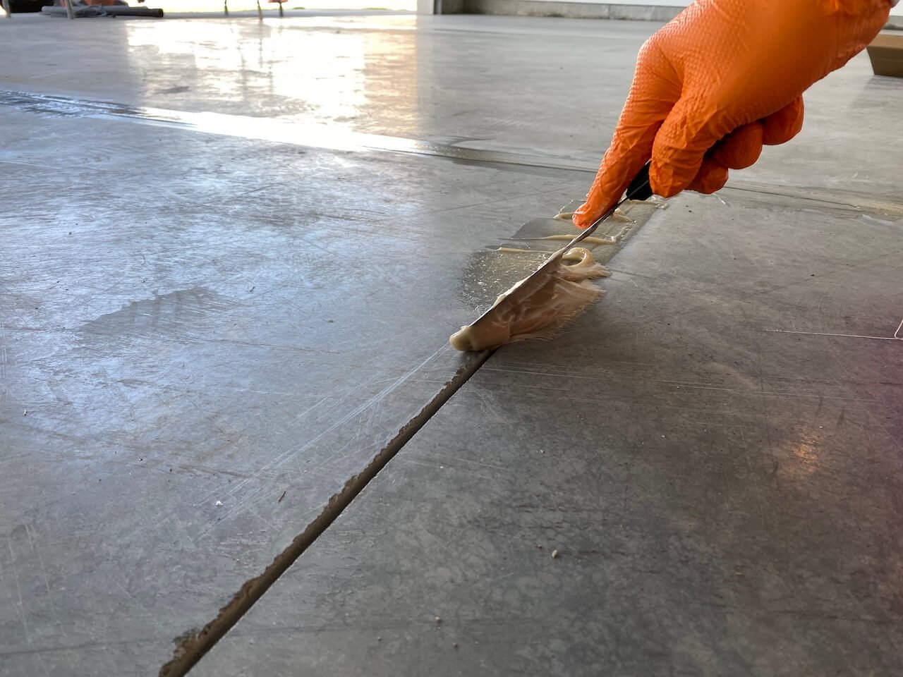 Person applying sealant to concrete floor crack with putty knife, wearing orange glove.