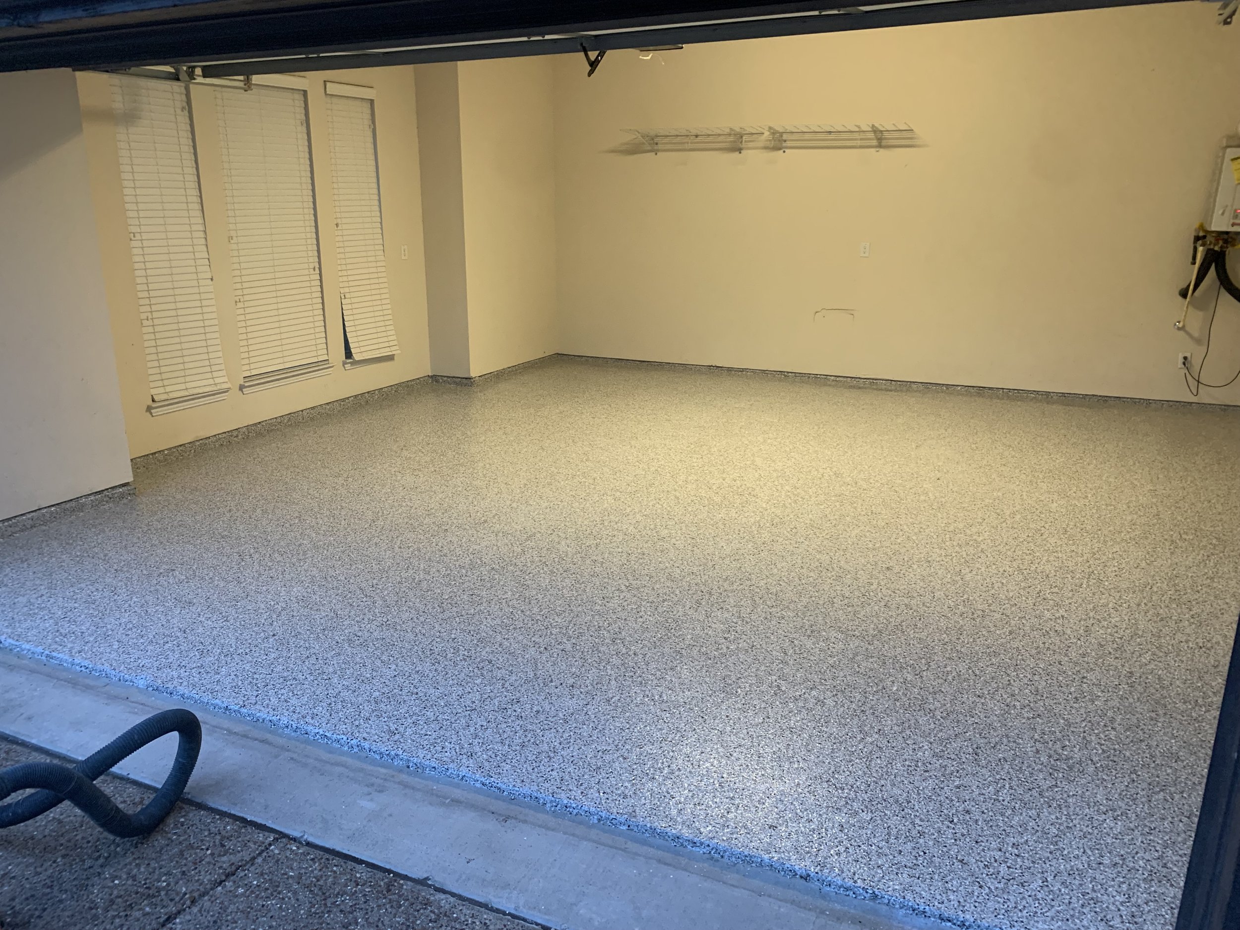 Empty garage with epoxy-coated floor and white walls, featuring large windows with blinds and an overhead storage rack.