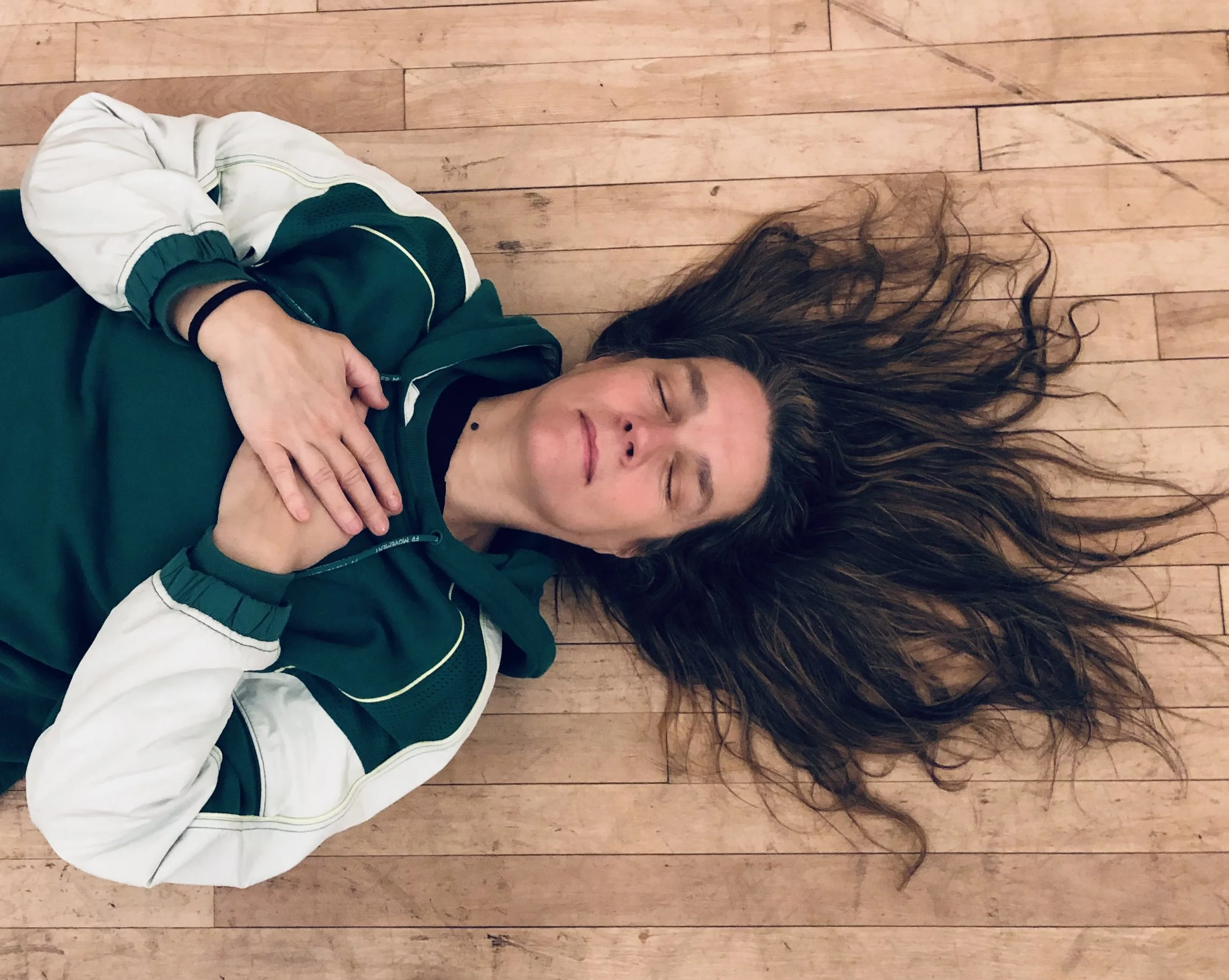 A woman lying on a wooden floor, eyes closed, with her long hair spread out around her head, wearing a green and white athletic jacket.