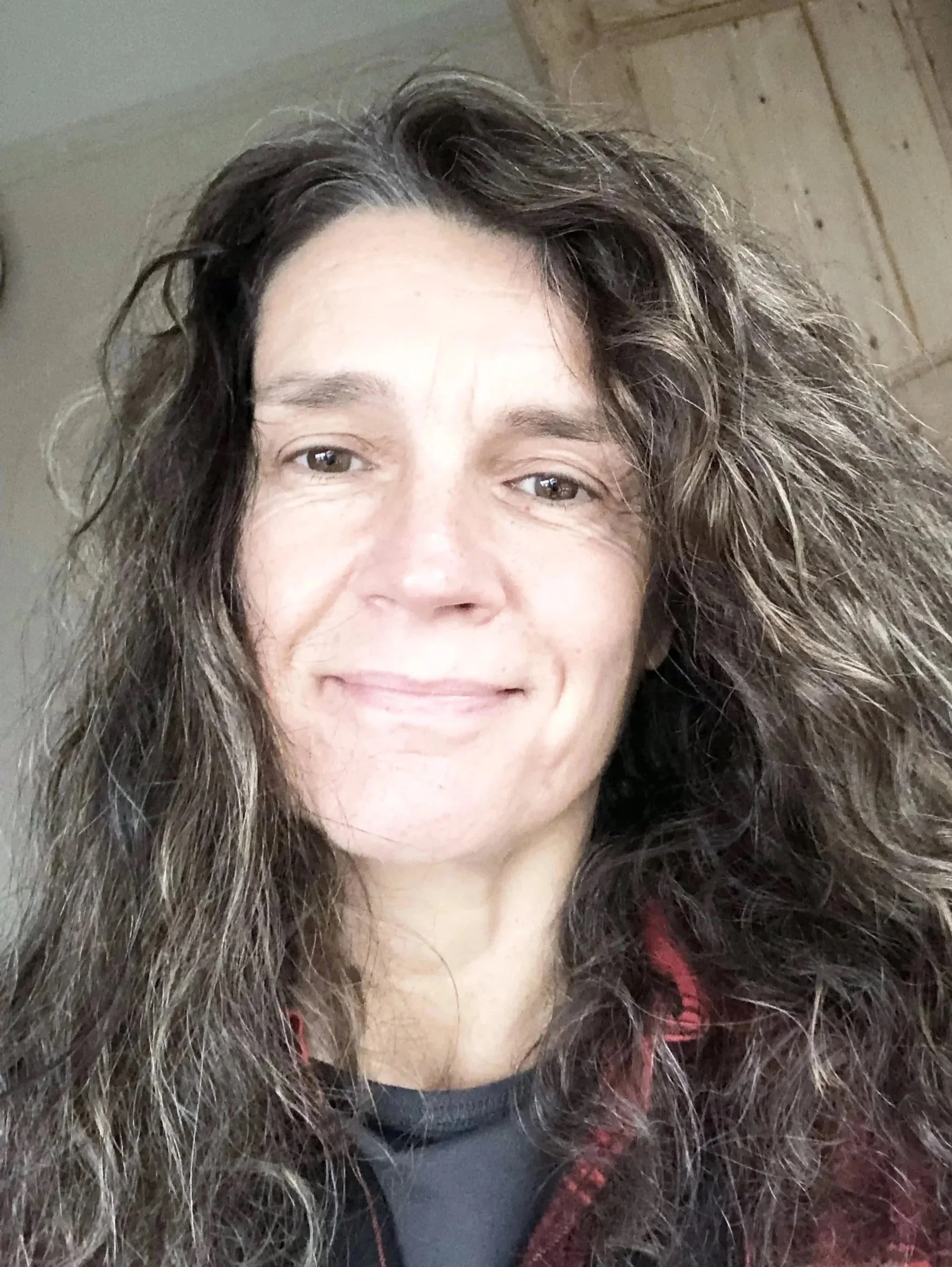 Close-up of a woman with long, curly brown hair, smiling softly, indoors with a wooden ceiling in the background.