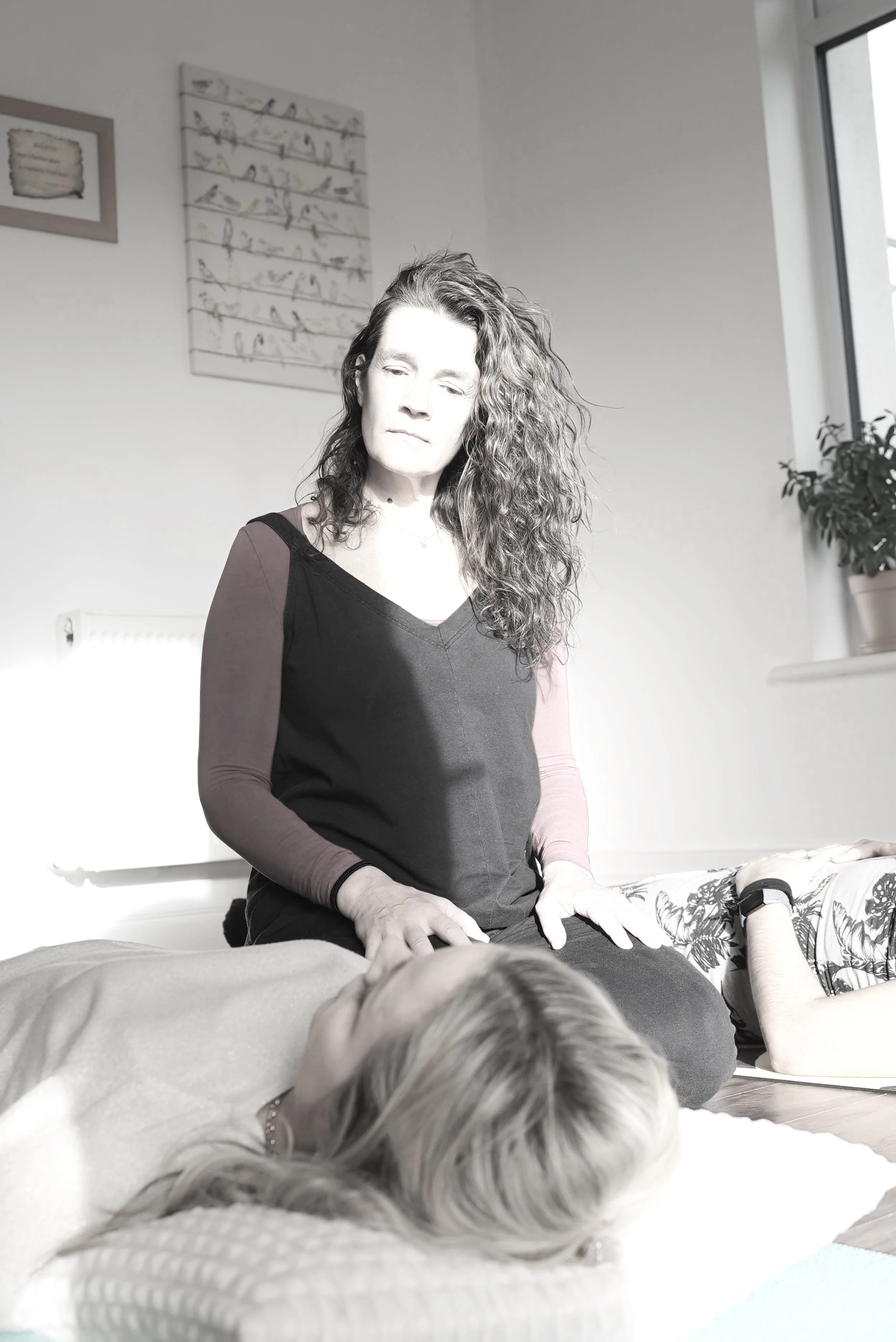 A person with curly hair is performing a healing or relaxation practice on another person lying down. The room is well-lit with minimal decoration, including framed art on the wall and a potted plant by the window.