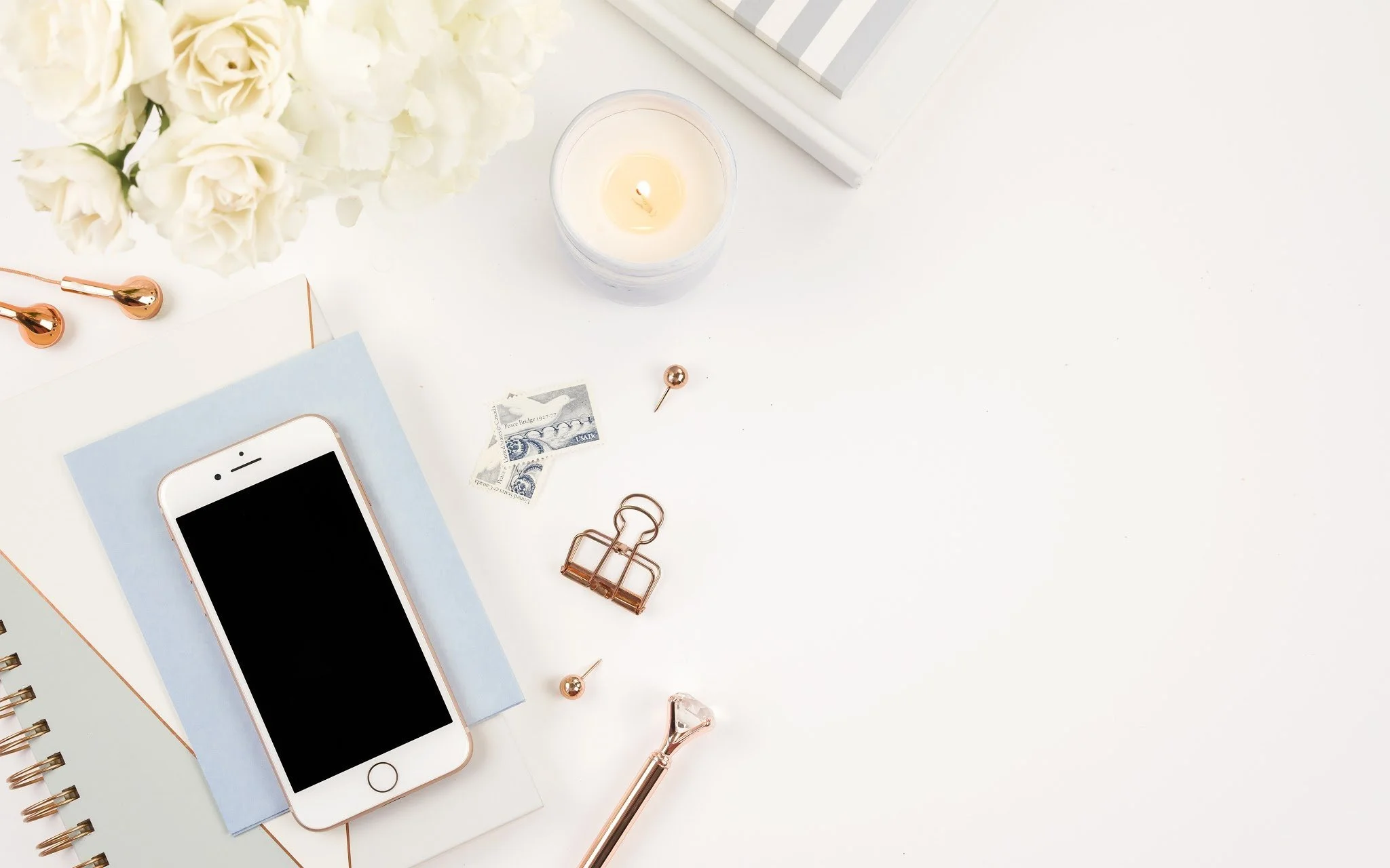 A white desk decorated with a bouquet of white roses, a lit candle in a glass holder, a smartphone with a black screen, notebooks, rose gold stationery, miniature dollar bills, and small decorative items.