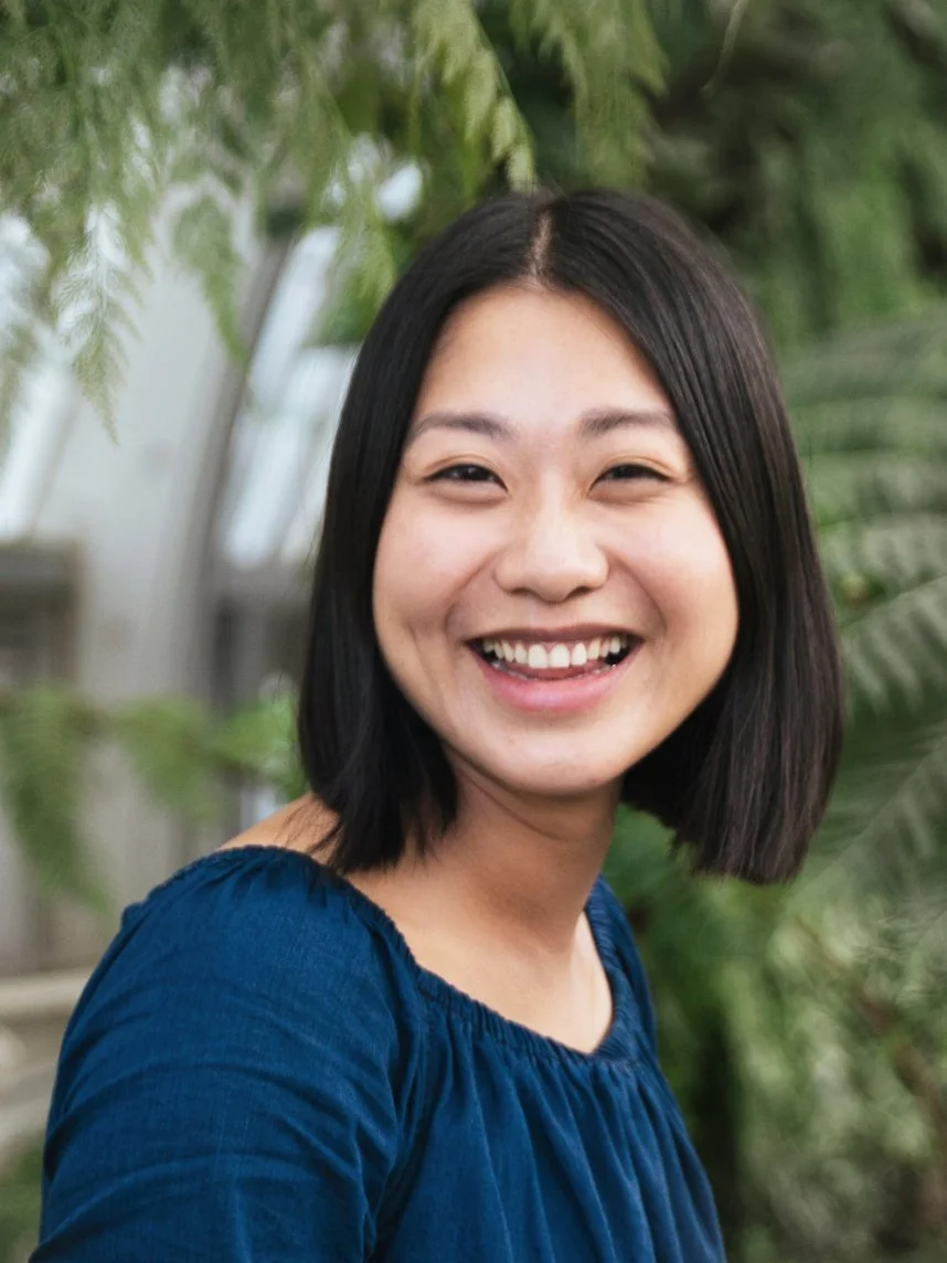 A smiling woman with short black hair wearing a blue top, standing outdoors with green foliage in the background.