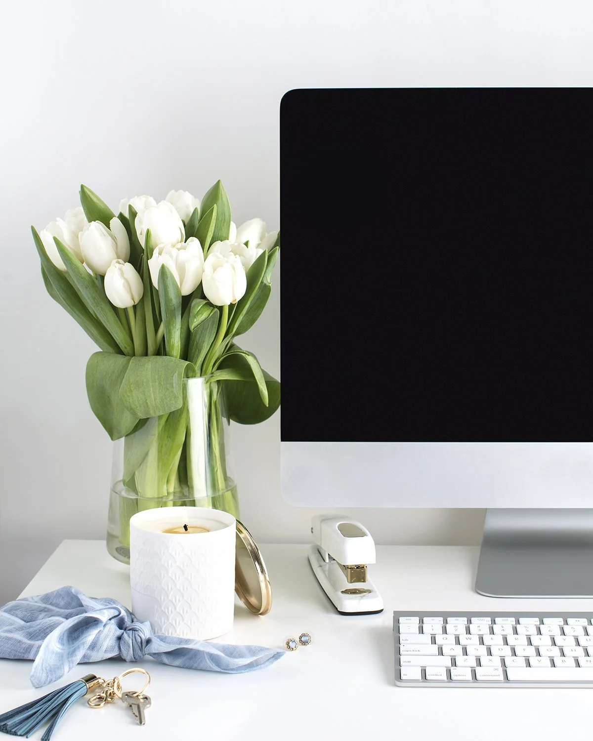 A desktop workspace with a bouquet of white tulips, a lit white candle, a stapler, a computer monitor, a keyboard, and various jewelry items on a white surface.