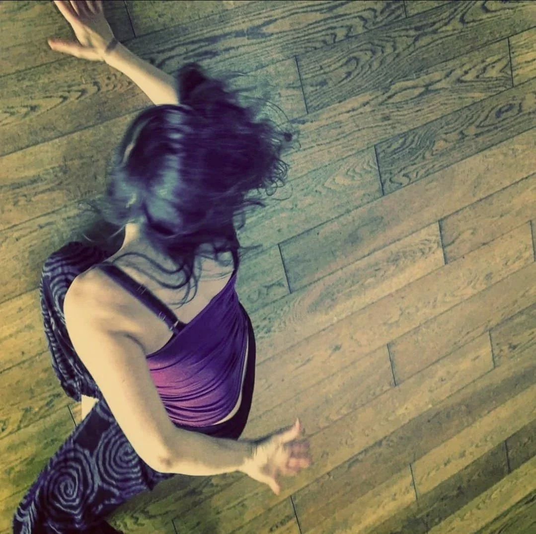 A woman with long dark hair dancing on a wooden floor, wearing a purple dress and patterned pants.