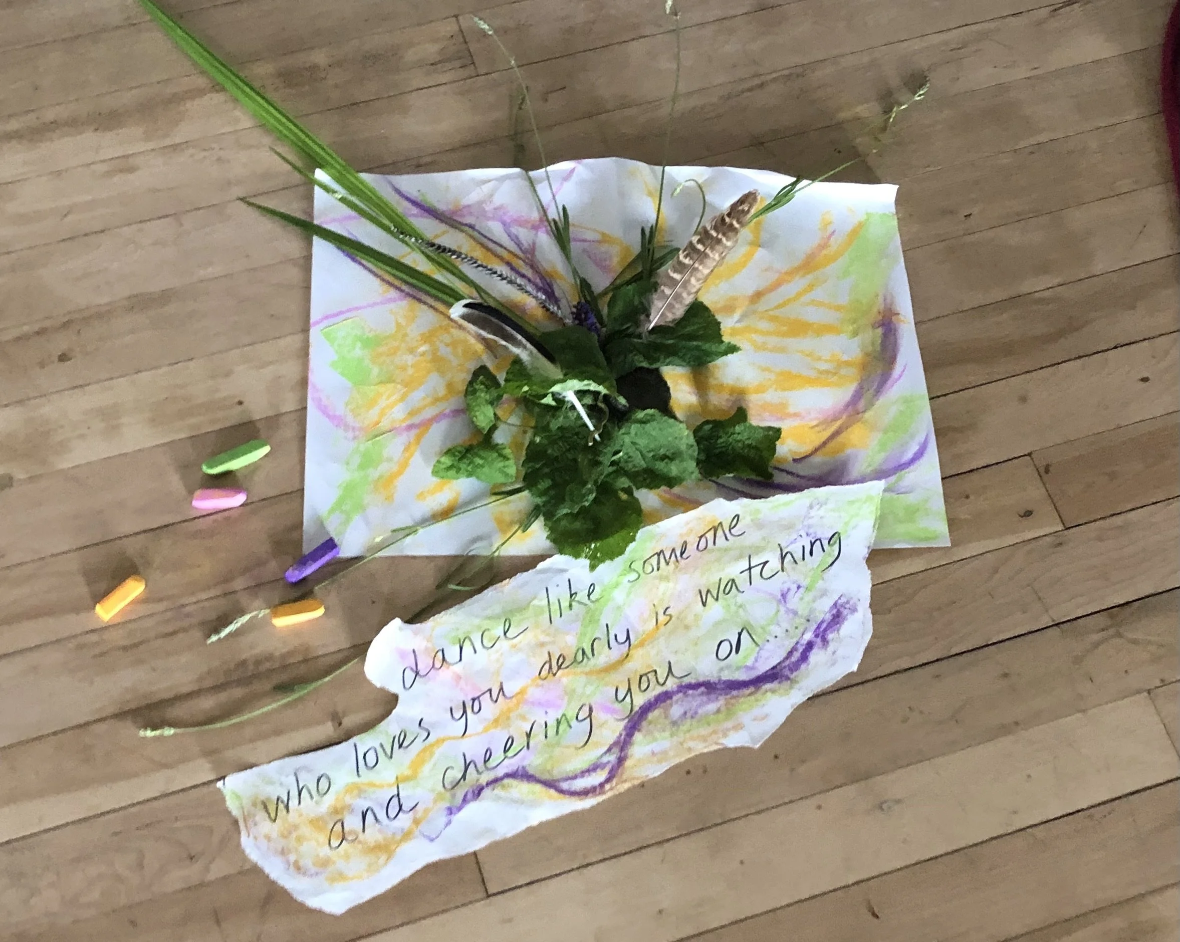 A handmade arrangement of flowers, feathers, and green leaves on a brightly colored painted paper with a handwritten note that says "dance like someone who loves you dearly is watching and cheering you on".