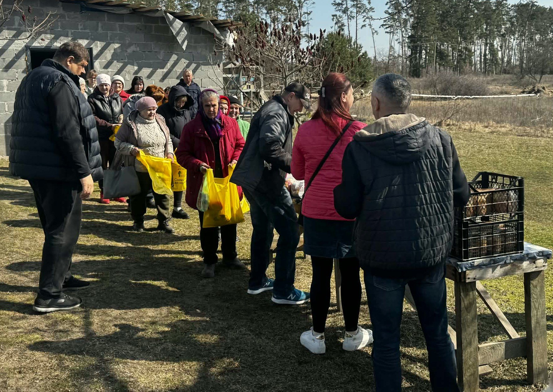 A queue of people stand outside a brick building, beside a table. they hold plastic bags which they are filling with food from the table.