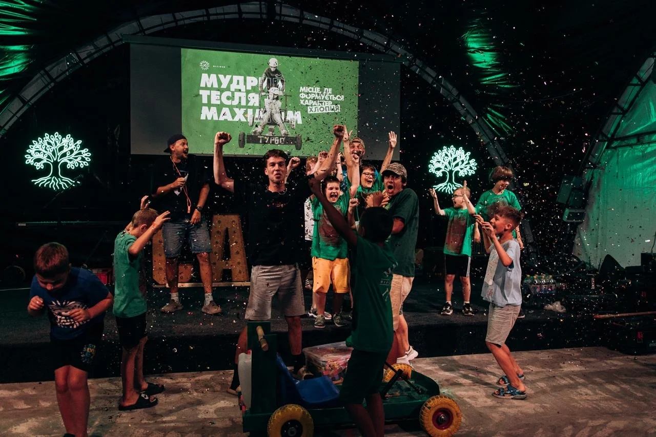 A group of boys wearing matching t shirts, with their hands in the air in celebration. They are laughing and jumping, in front of them is a small car they have constructed from wood.