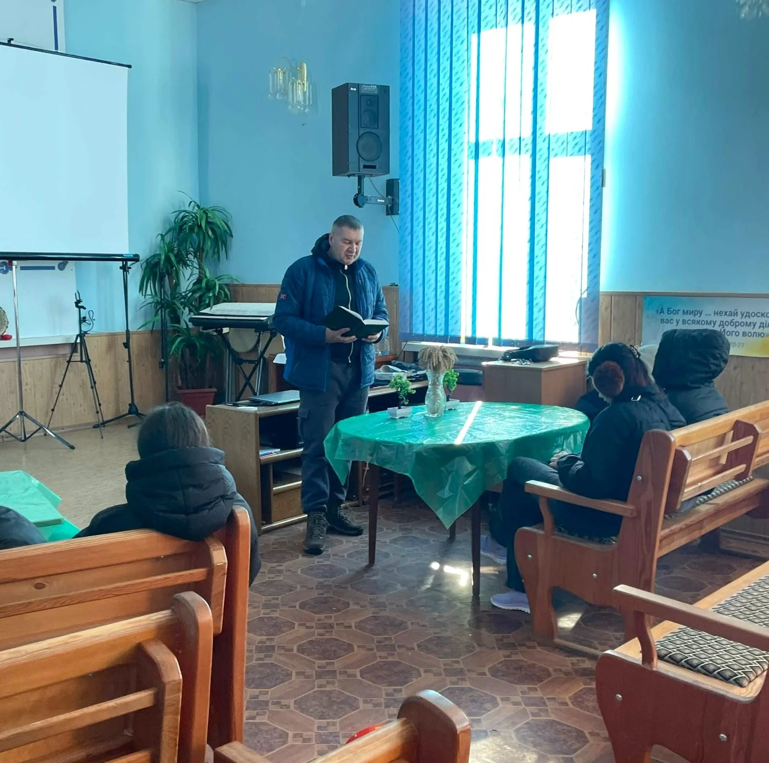 A man stands at the front of a church, holding a Bible. Some people are facing him, sitting on pews.