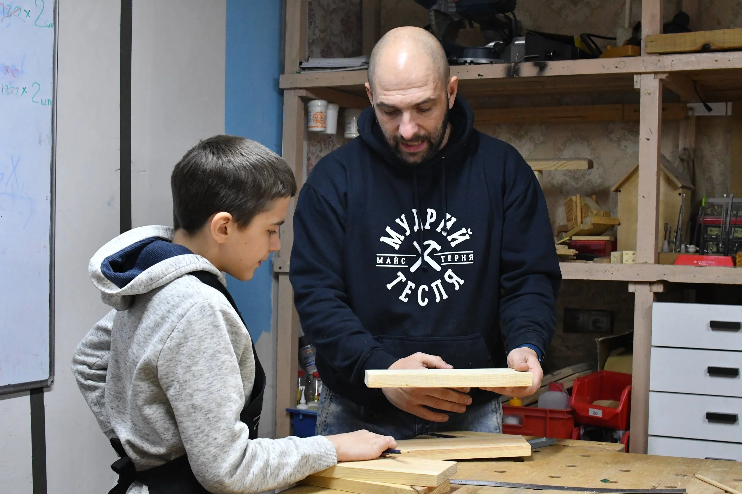 A man in a carpentry workshop is holding a piece of wood and showing a teenage boy where he should cut it.