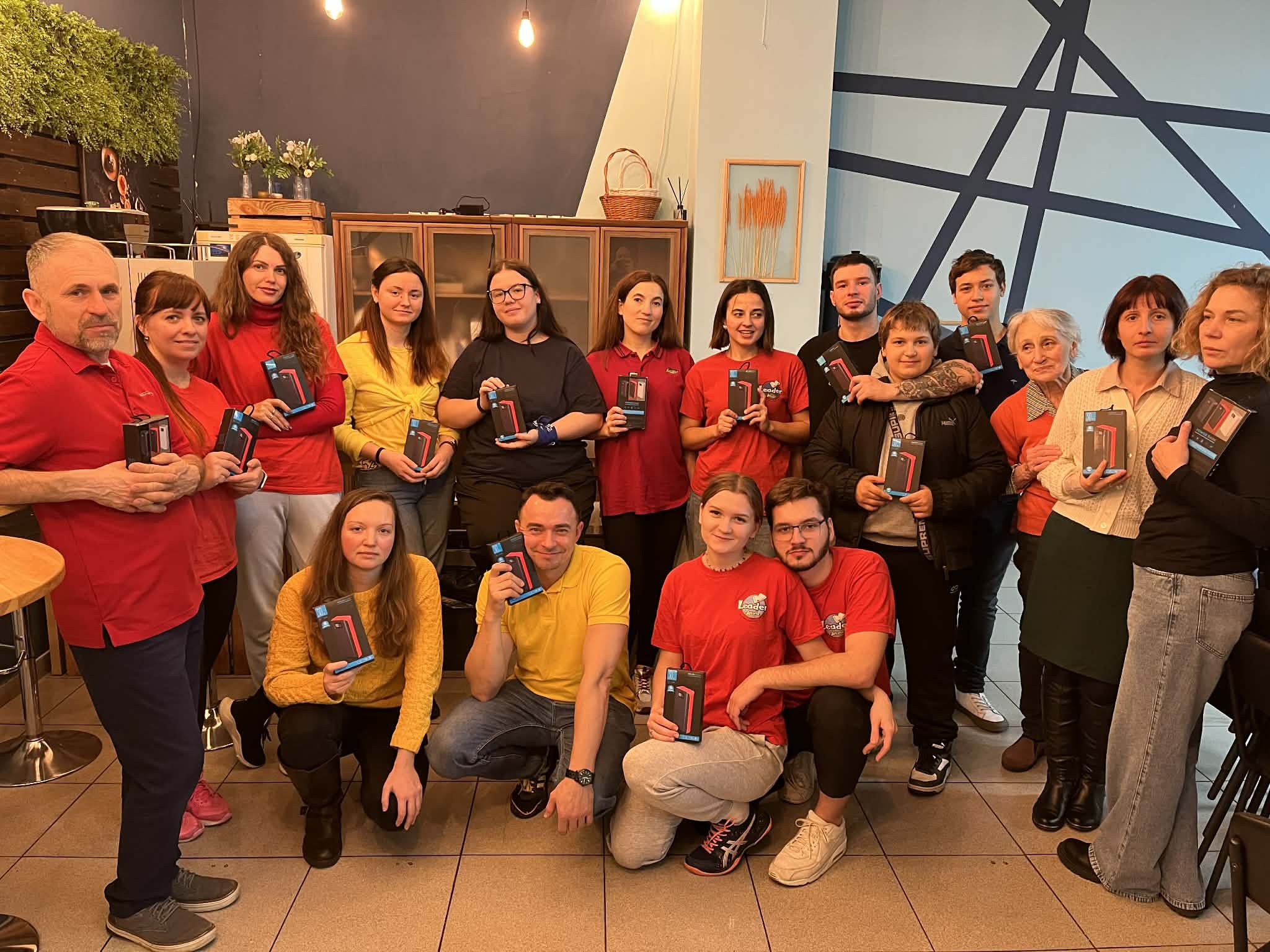 A group of men and women, all different ages, mostly wearing red t shirts. Some are standing in a semicircle and a few kneeling in front. Each person is holding a power bank for charging a mobile phone.