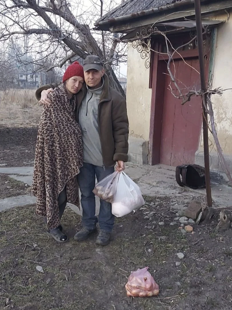 A man and woman standing in a front yard, in front of a house. He has his arm around her shoulders and she is wrapped in a blanket. They look sad. He is holding 2 plastic bags with food in.