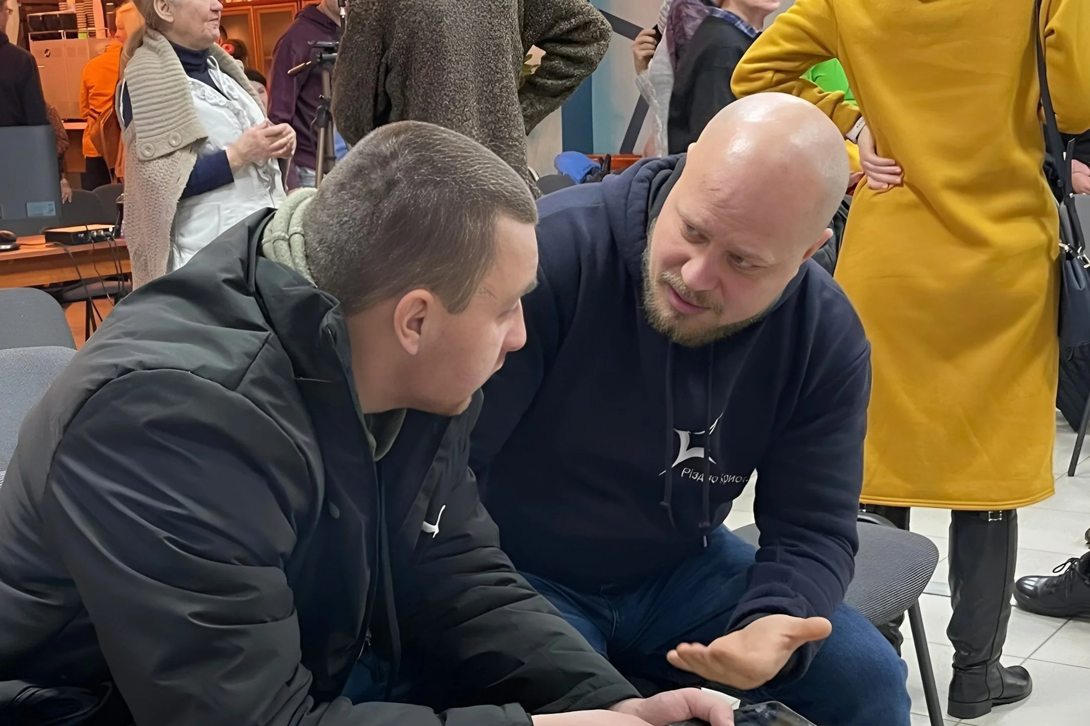 Two men are sitting next to each other, deep in conversation. A few other people are standing around behind them.