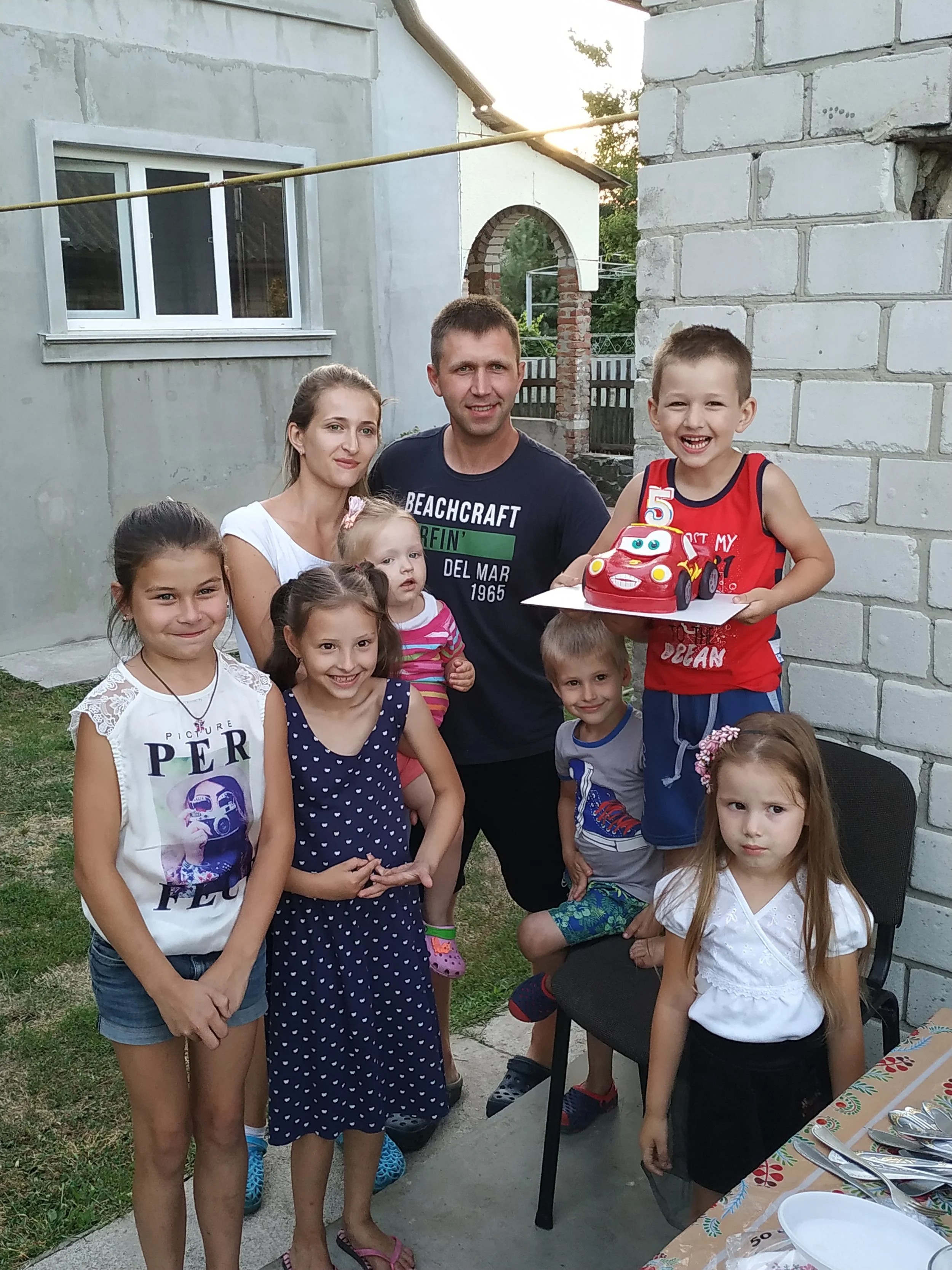 A family group are celebrating a boy's birthday with a cake shaped like a red racing car. There are two parents, five children and a baby in her mother's arms. They are all smiling.
