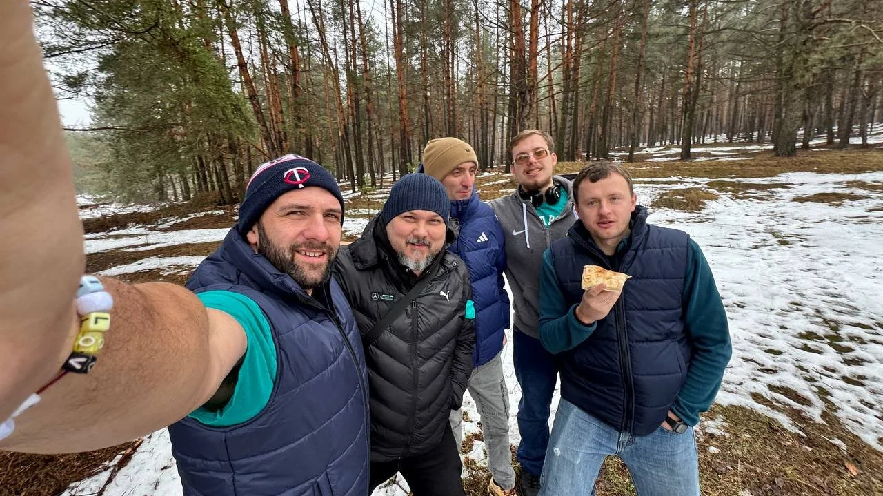 Five men are standing together outside in a forest clearing, with snow on the ground. They are wearing winter clothes, looking at the camera and smiling.
