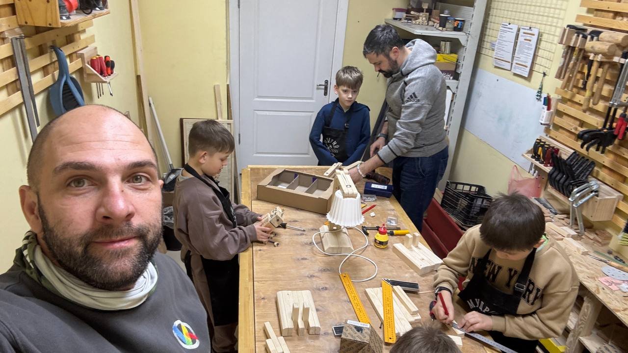 A man is looking at the camera and behind him three boys are doing activities at a woodworking table, assisted by another man. Around them are tools for doing carpentry.