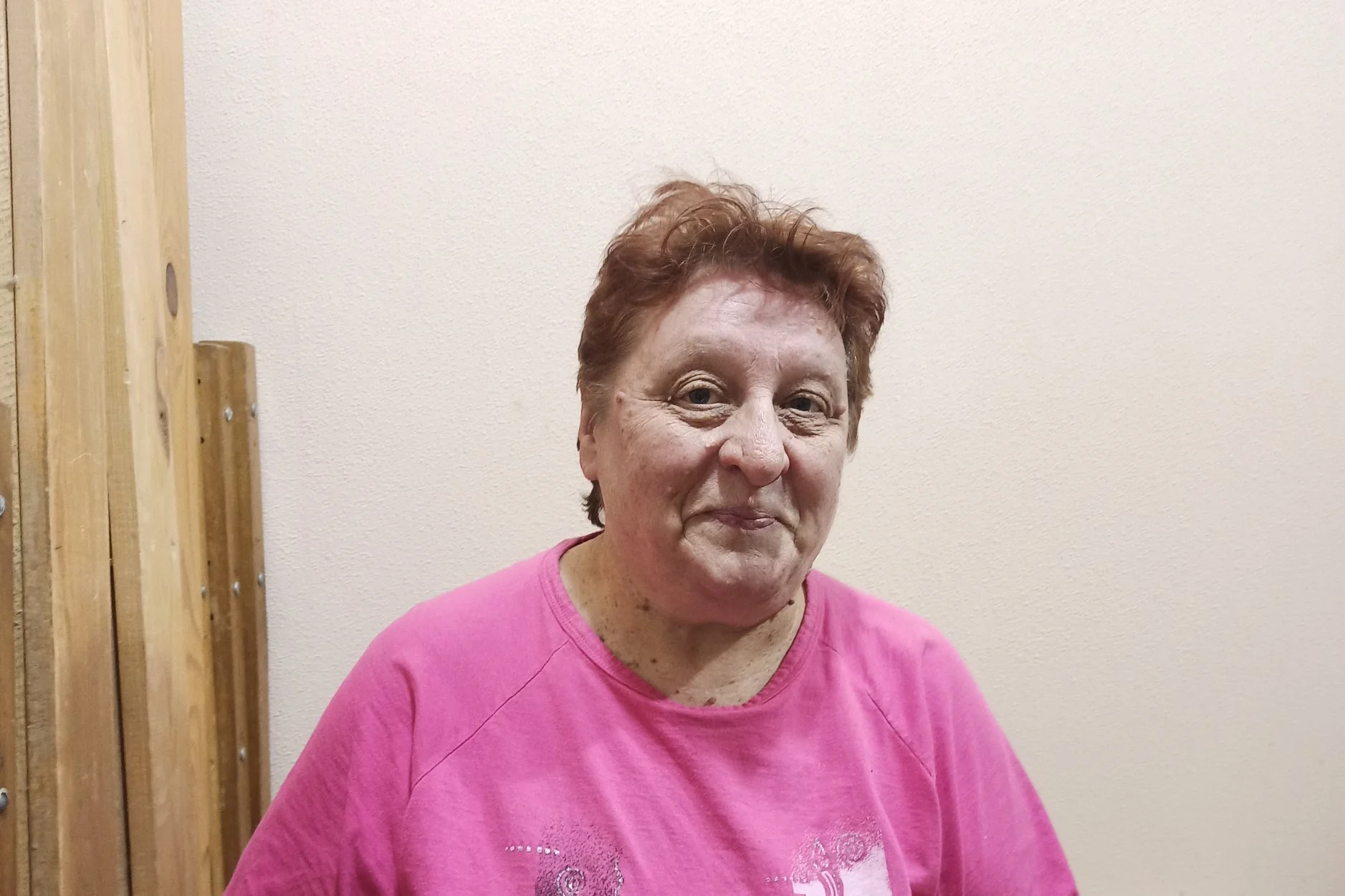 A lady stands in a room smiling at the camera, wearing a pink t-shirt