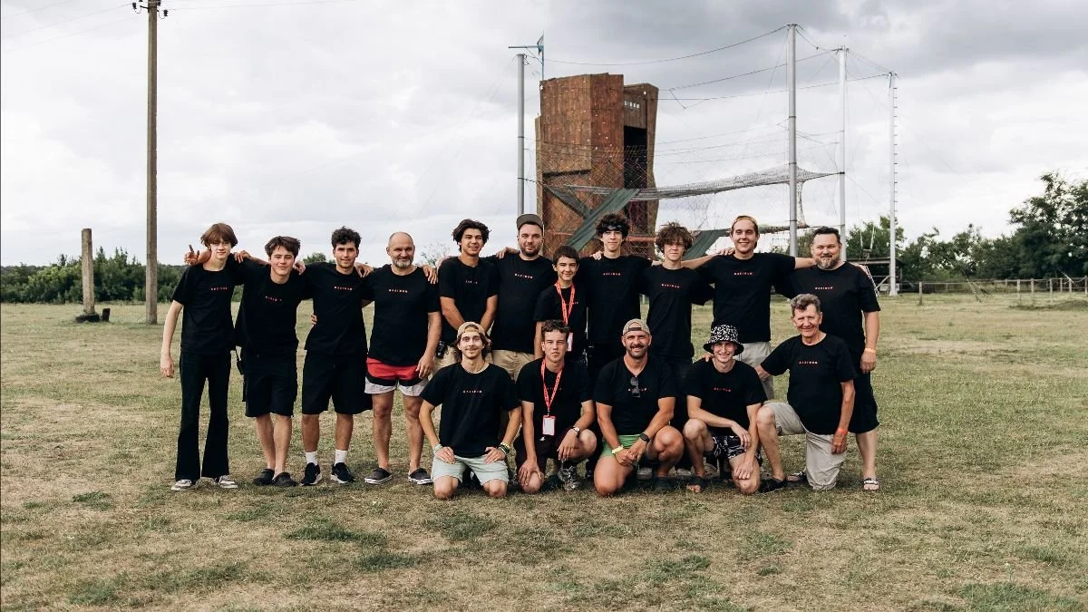 A group of teenagers and older men, posing for a picture, some standing with arms around shoulders, others kneeling in front. They are in a large field, with a climbing wall behind them.