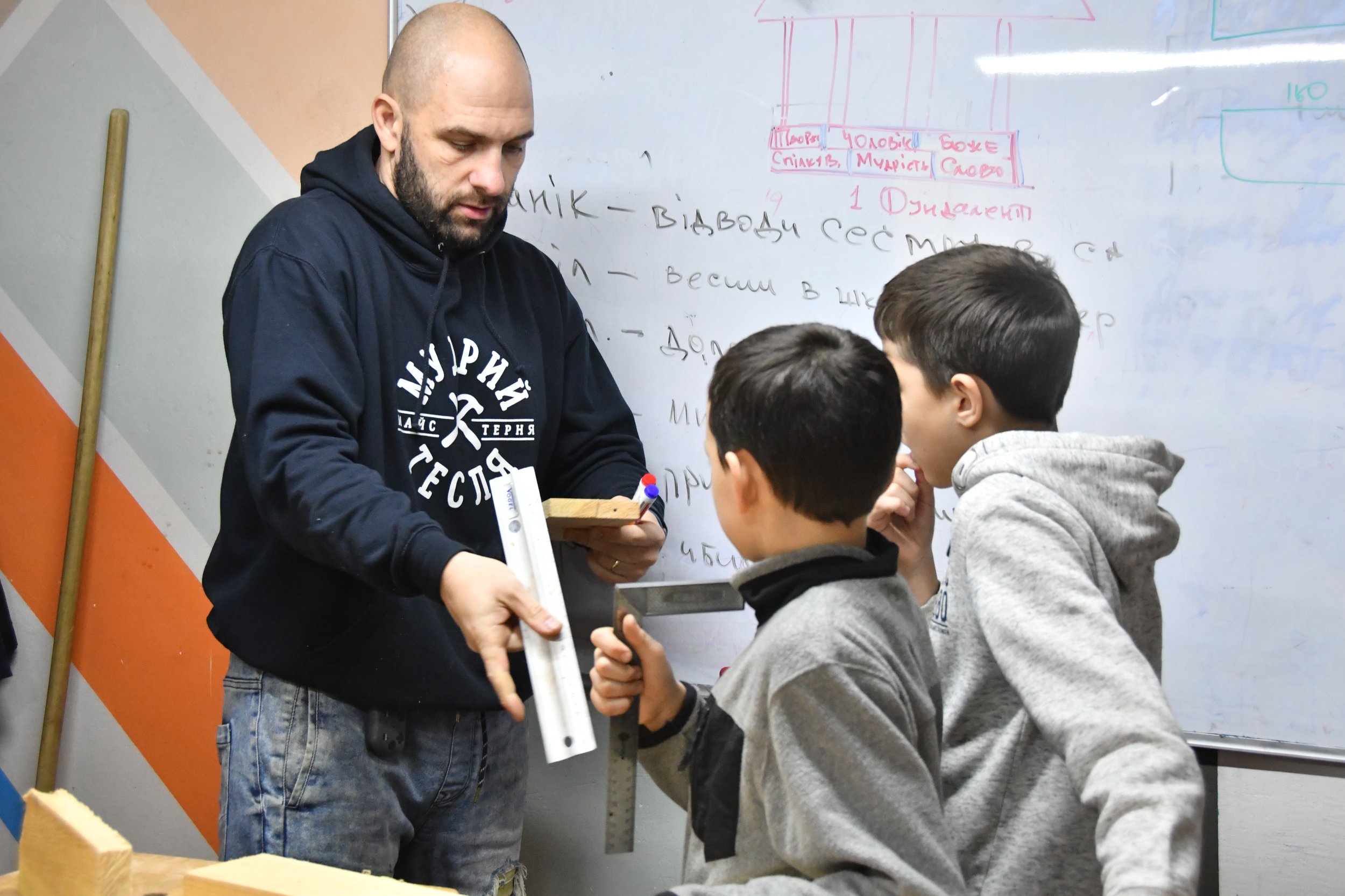 A man is holding a piece of wood and a metal ruler. He is demonstrating to two young boys how to measure the wood.