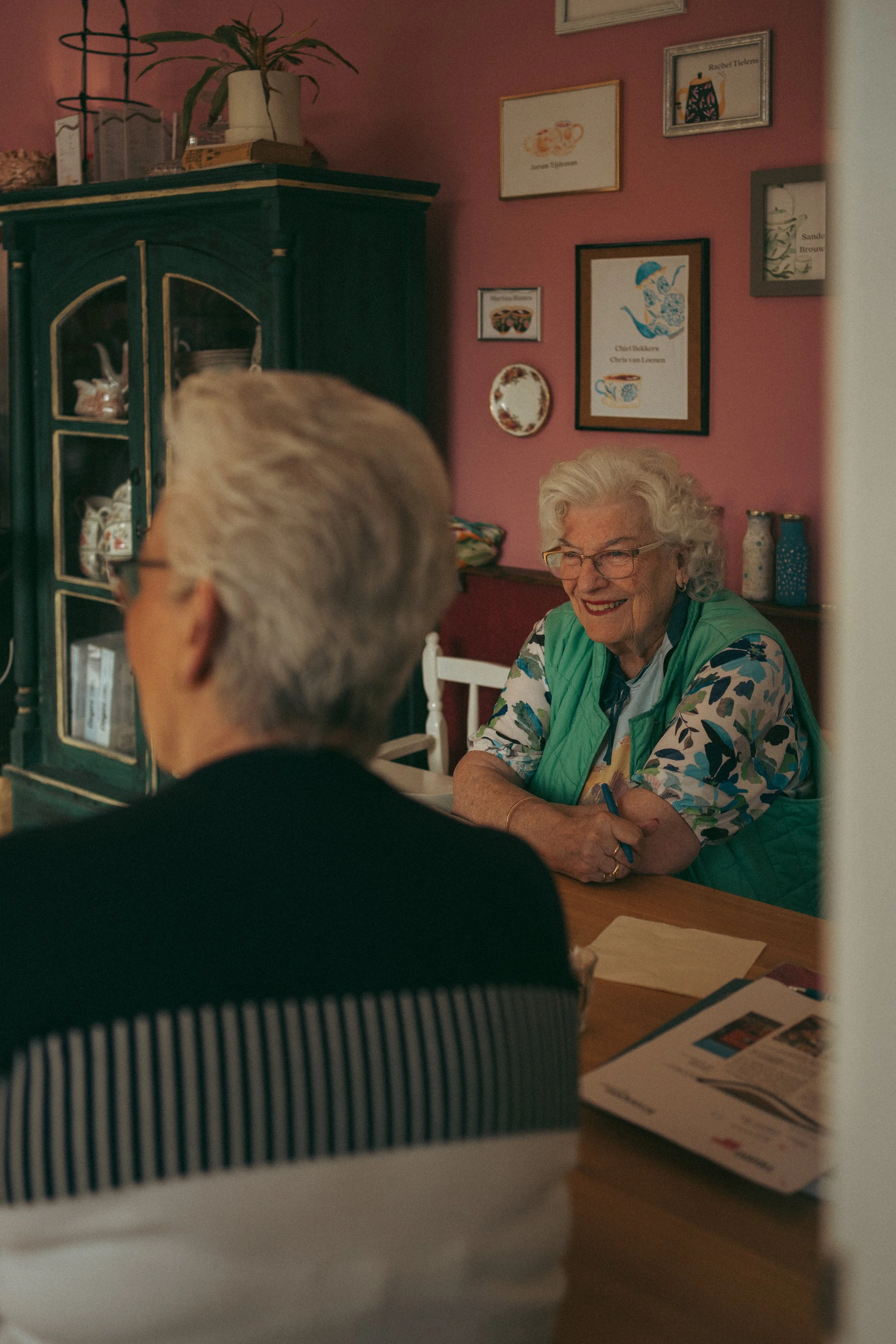 Een oudere vrouw lacht terwijl ze met een man aan een eettafel zit, in een gezellige keuken met roze muren en diverse schilderijen aan de muur.