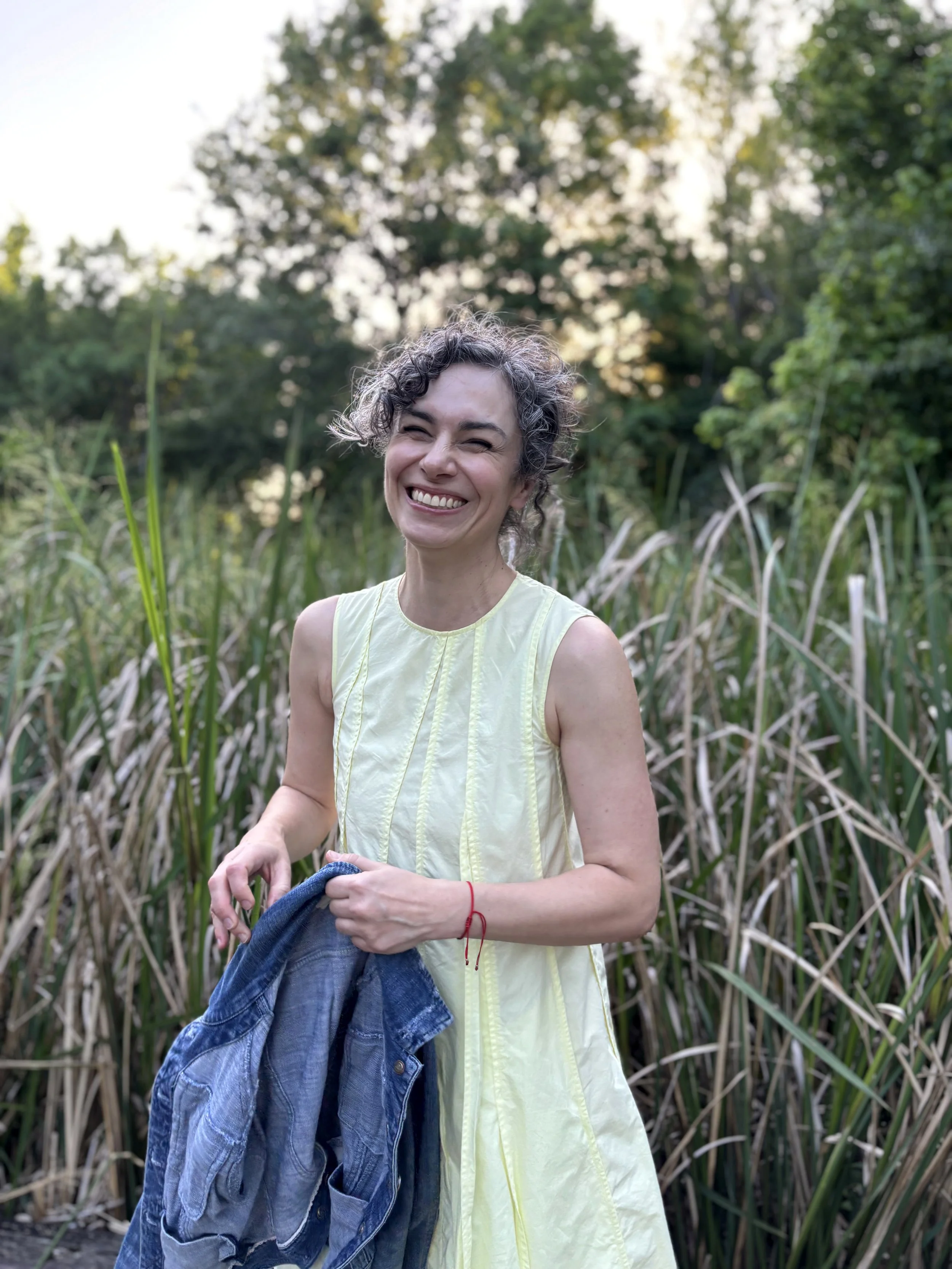 A smiling woman with curly hair in a sleeveless yellow dress holding a denim jacket, standing outdoors in a natural setting with tall grass and trees in the background.