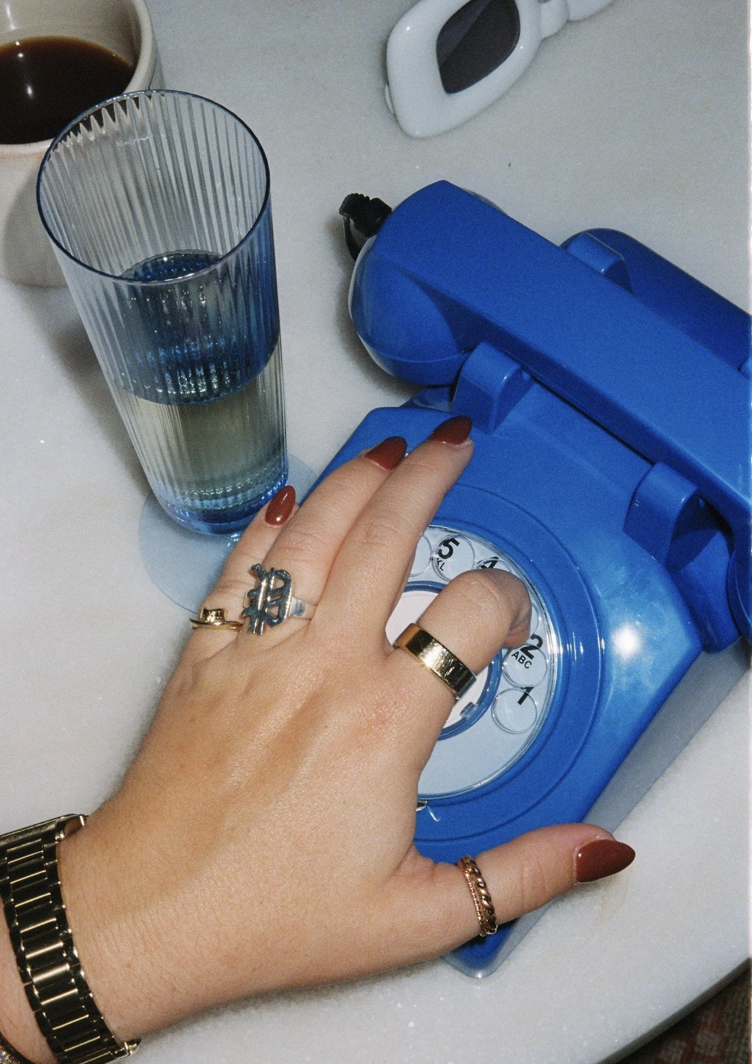 A hand with rings and a watch pressing the button on a blue rotary phone, with a glass of water and a cup of coffee nearby on a white surface.