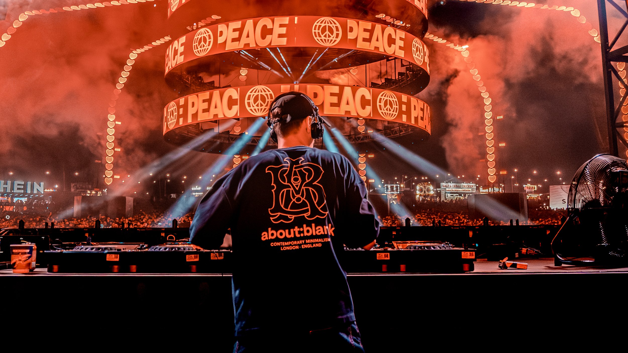 A DJ performing at a large music event with orange lighting, smoke effects, and screens displaying the word 'PEACE' along with peace symbols.