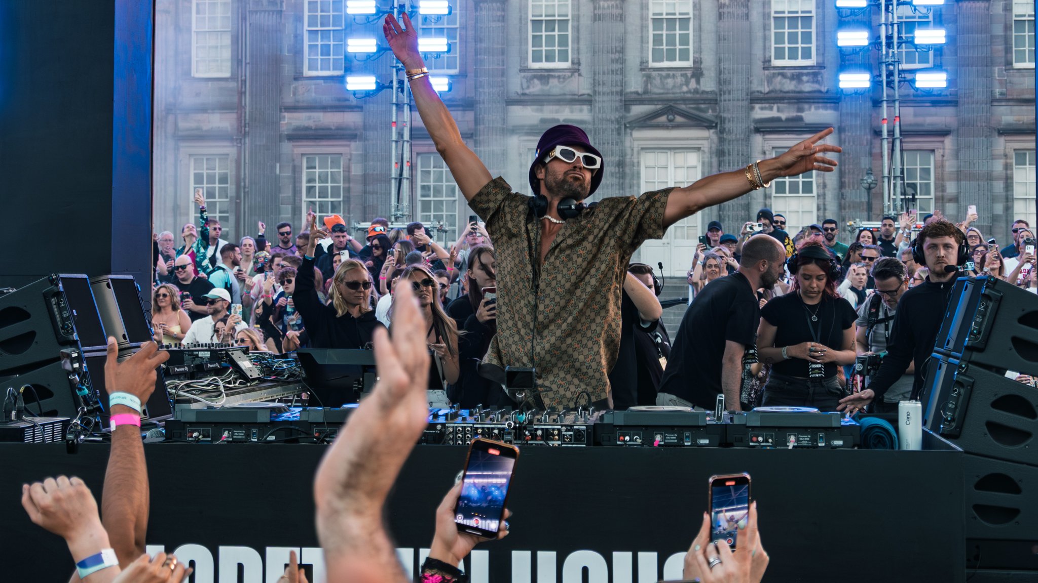 A DJ with sunglasses and a patterned shirt is performing at an outdoor music event, surrounded by a crowd of people taking photos and enjoying the music, with a building and blue lights in the background.
