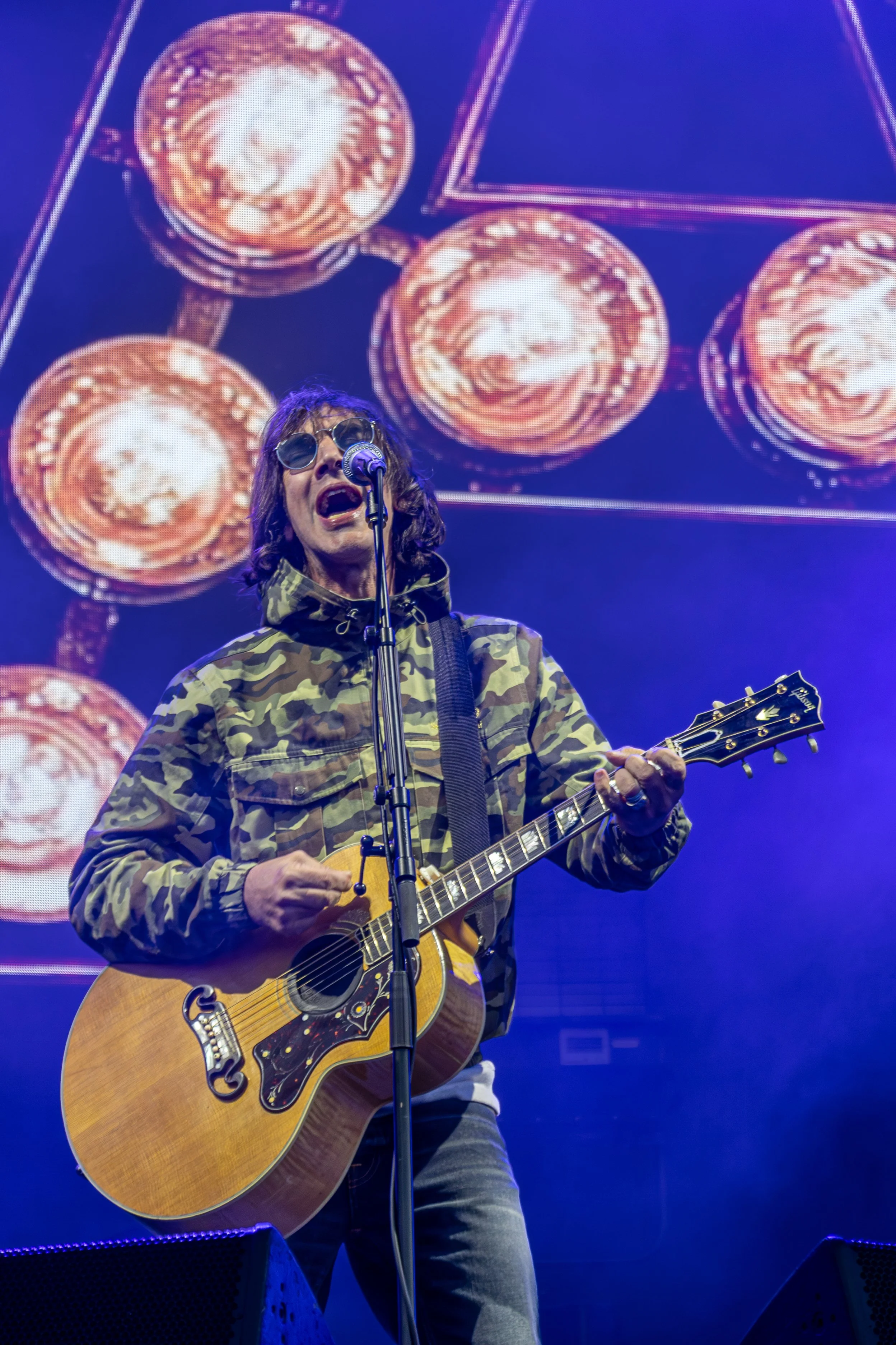 A male musician with long hair, wearing dark sunglasses and a camouflage jacket, singing into a microphone and playing an acoustic guitar on stage. A large screen behind him displays a graphic with circular shapes.