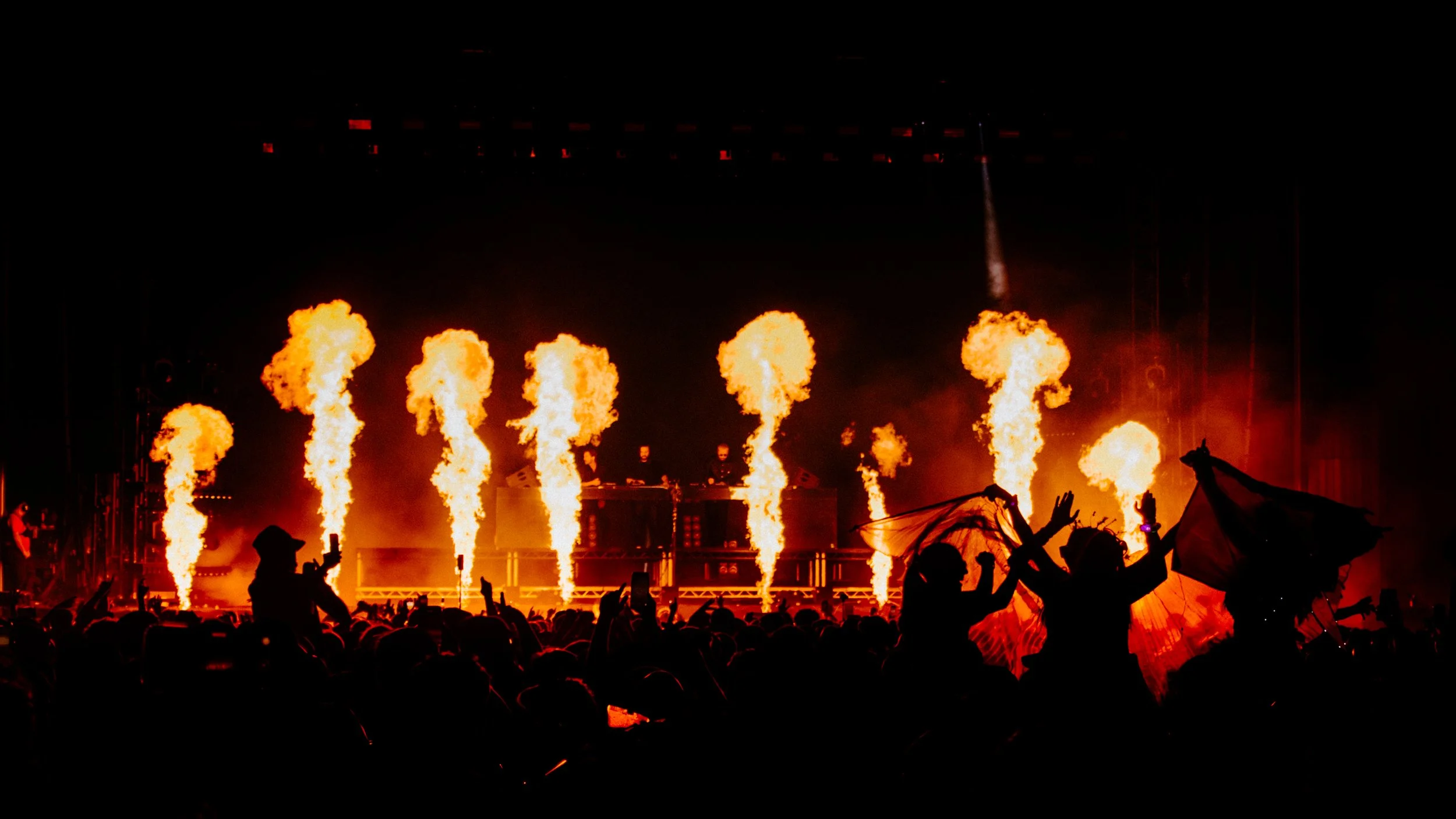 Crowd at a nighttime concert with flames and fire effects on stage, two DJs or performers at the center, and fireworks in the background.