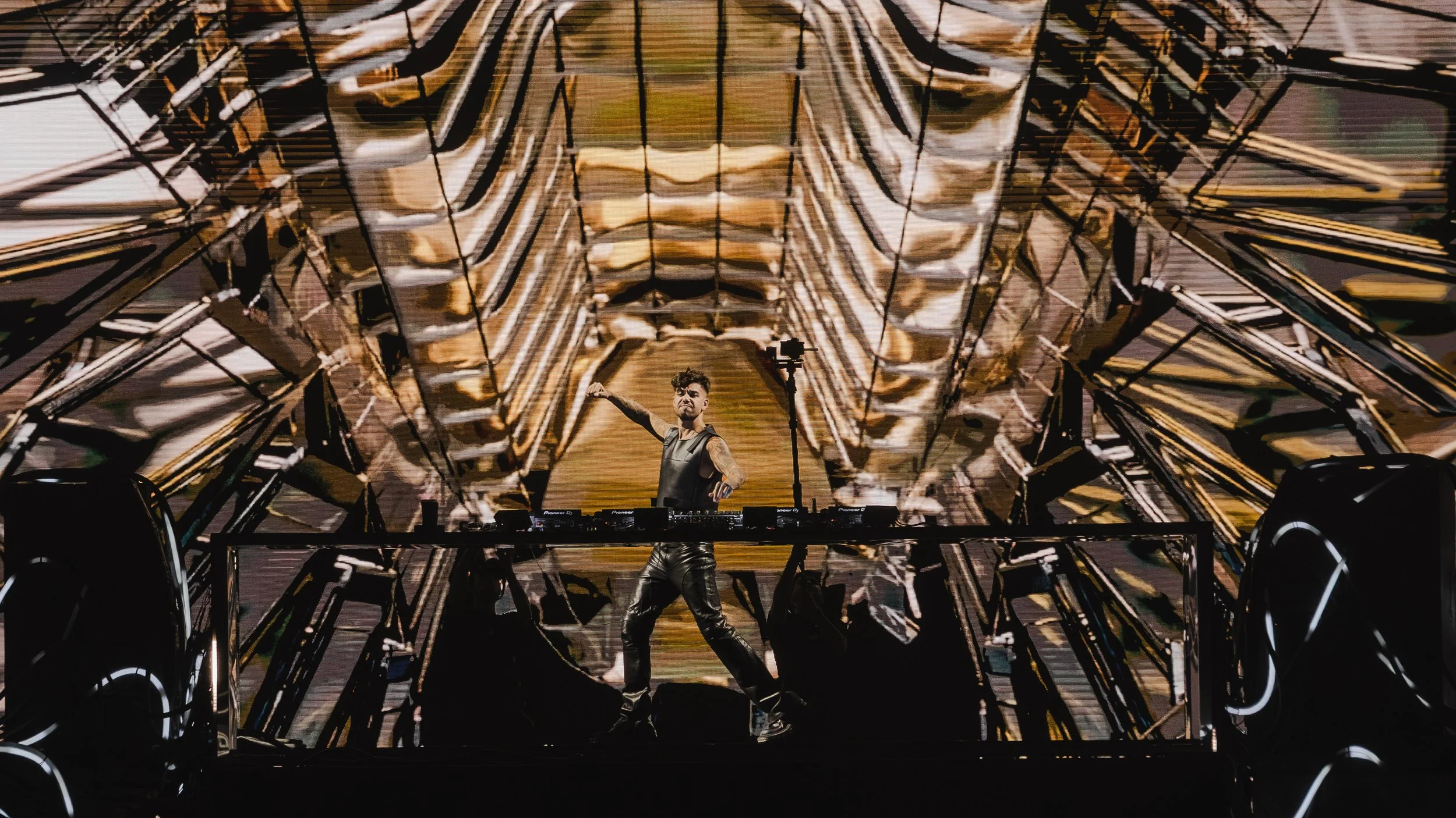A DJ performs on stage with a large, complex gold and black geometric structure behind him, featuring a screen displaying abstract visuals in warm tones.