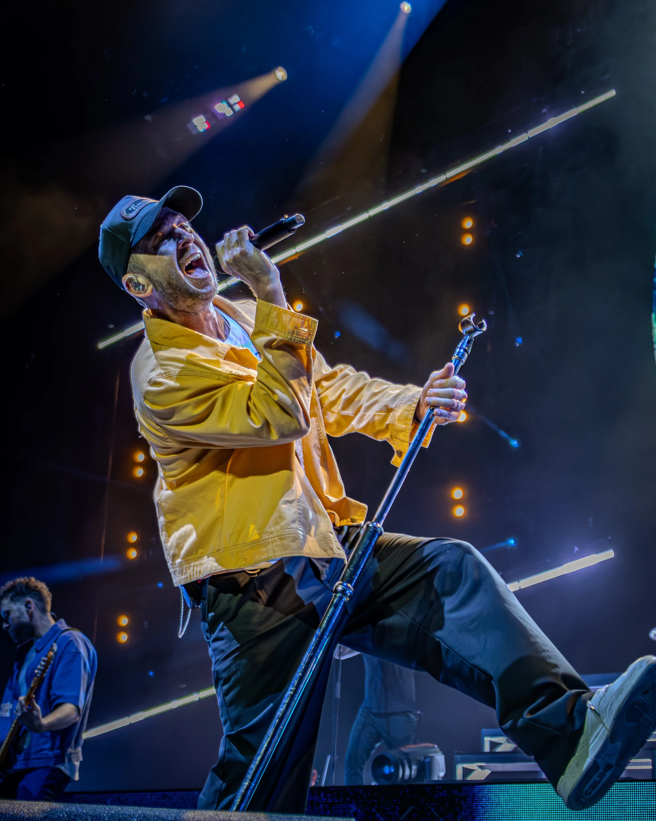 Performer passionately singing into microphone on stage during concert, wearing yellow jacket and dark pants, with stage lights and audience in background.
