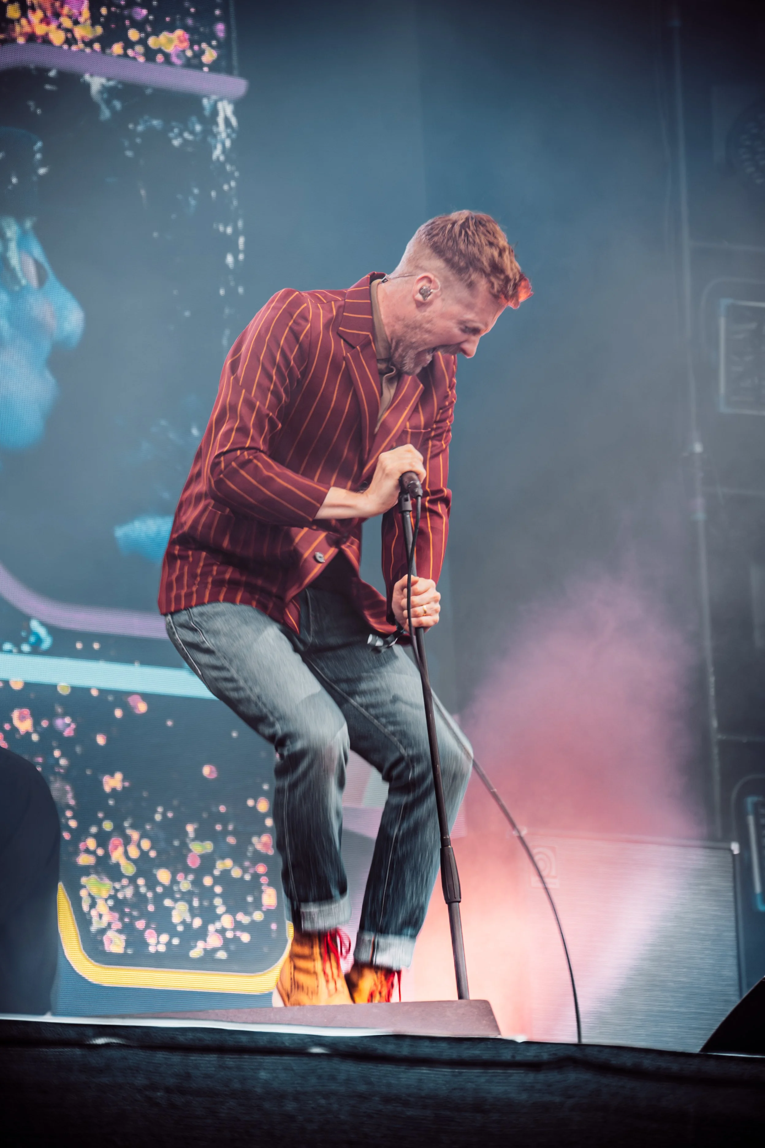 A man with short, reddish hair and a beard, wearing a brown and orange striped blazer, untied on the ground while singing into a microphone on stage.